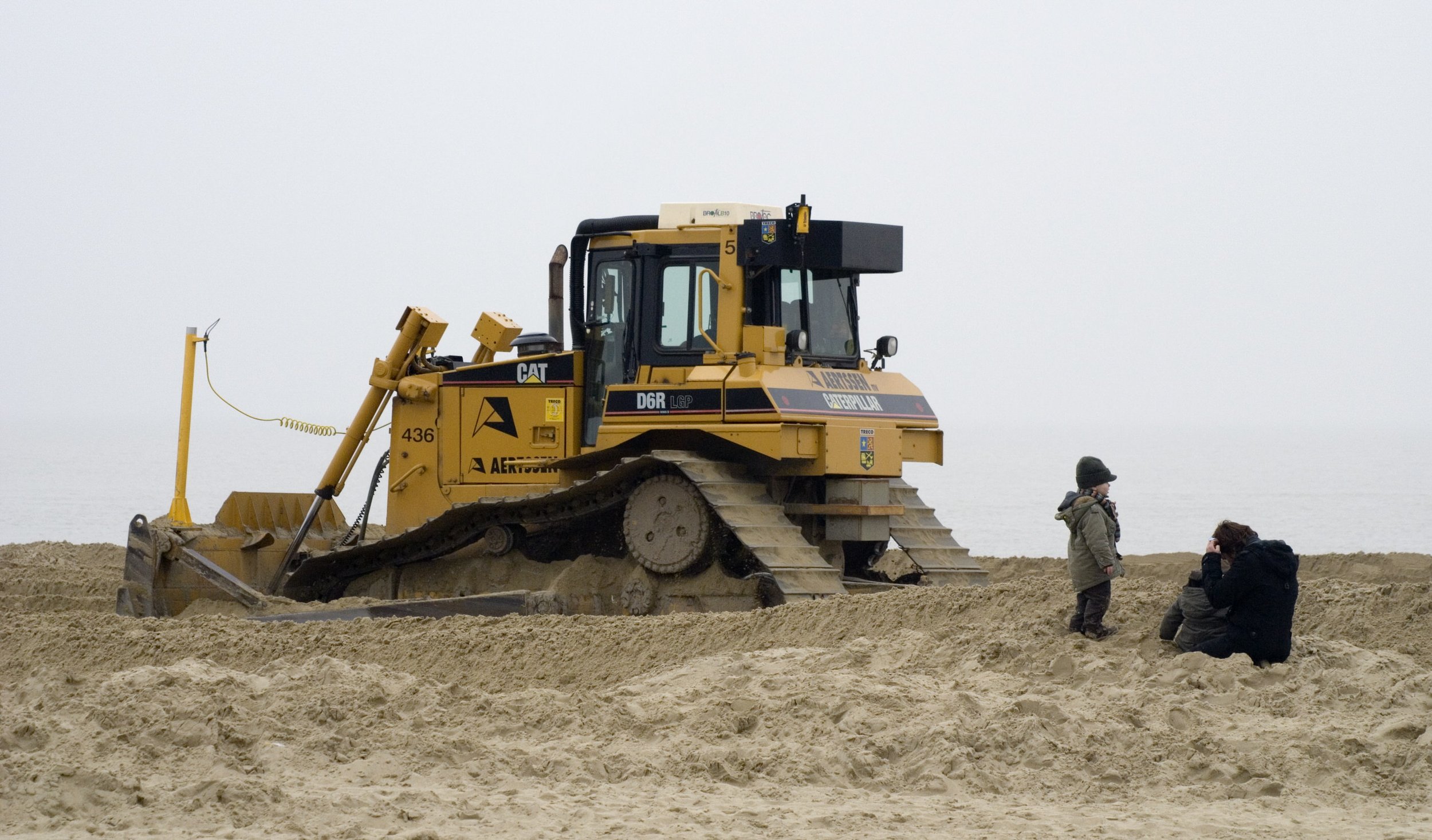 A yellow Caterpillar bulldozer working on a sandy beach with a person taking photos of two children playing nearby.