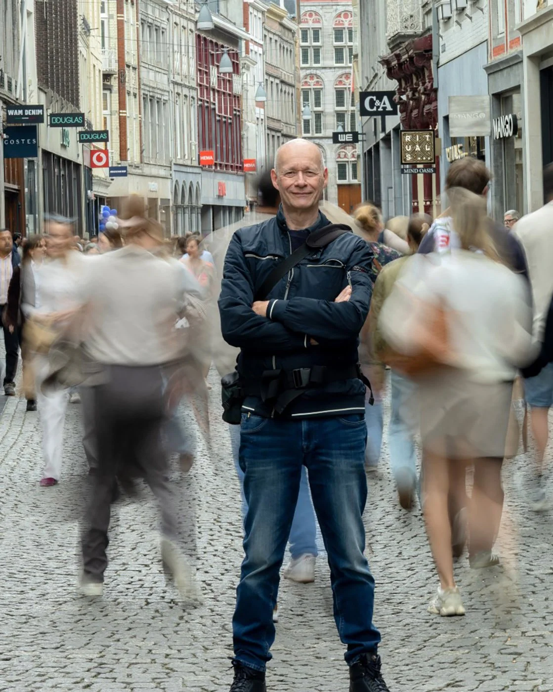 A man with a bald head, wearing a dark jacket and jeans, stands with arms crossed in the middle of a busy pedestrian street. Blurred people walk quickly around him, and tall historic buildings line the street, which is paved with cobblestones.