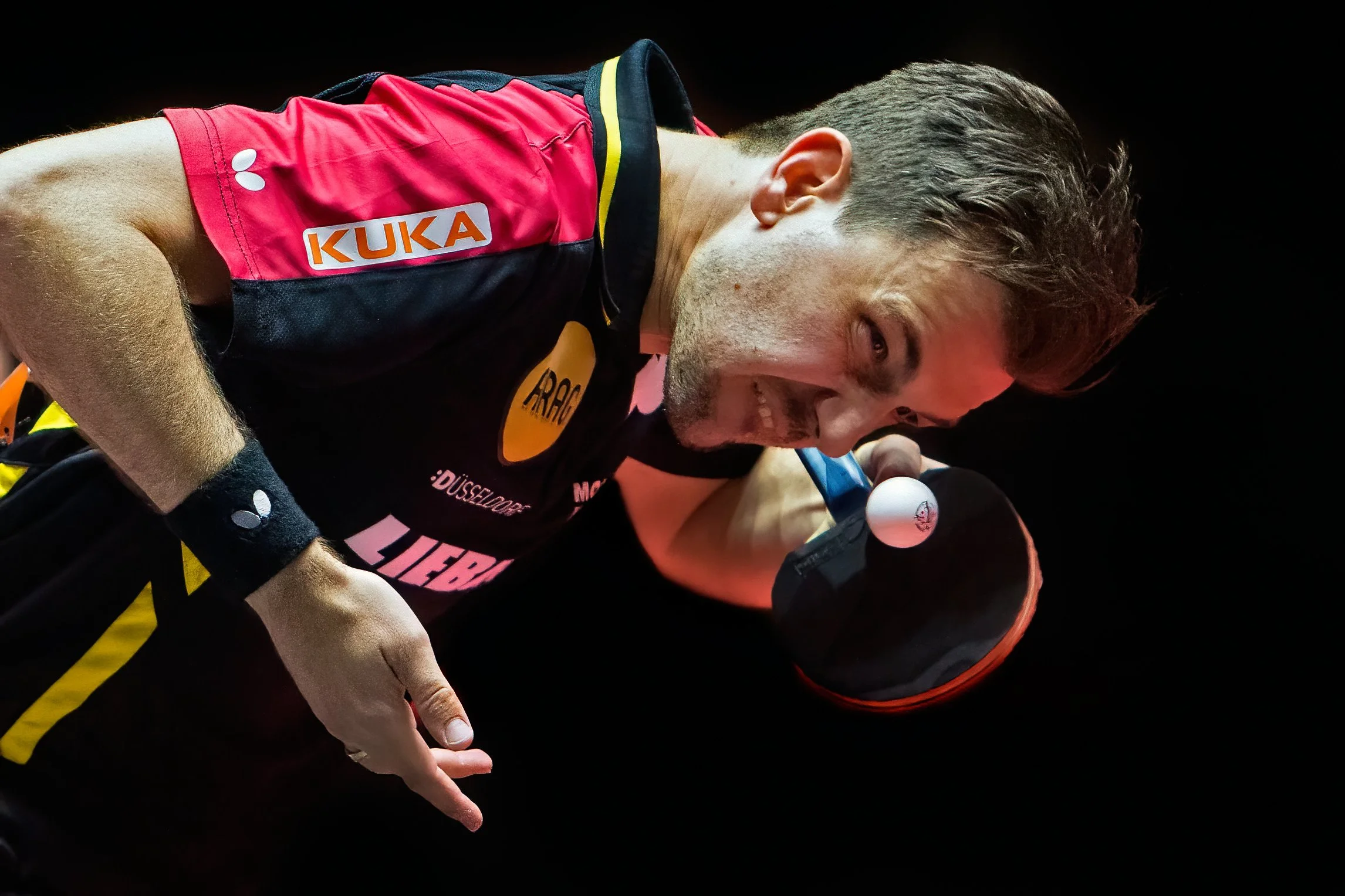 A male athlete in a pink and black sports uniform with sponsor logos, smiling and holding a ping pong paddle against a dark background.