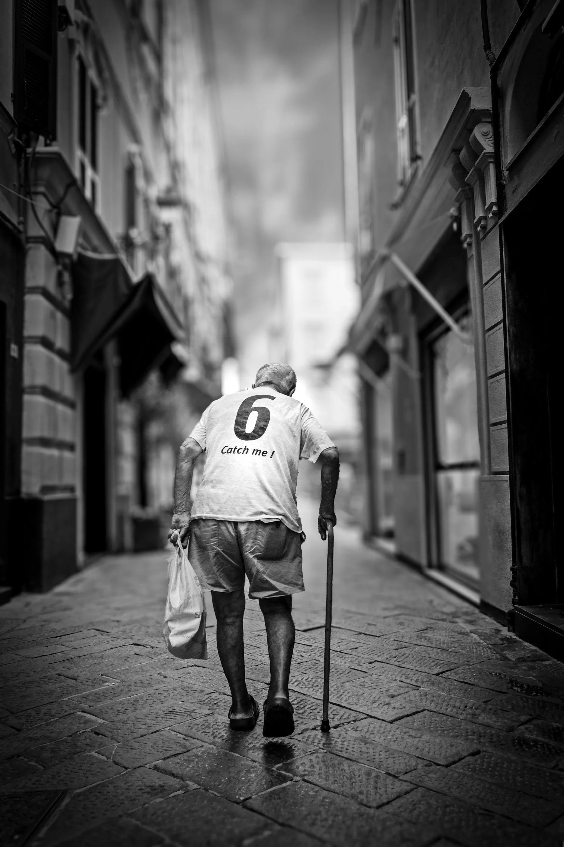 An elderly man with a walking stick, wearing a t-shirt with the number 6 and the phrase 'Catch me!' on the back, walking on a cobblestone street. He is carrying a plastic bag and holding a cane, with his back turned toward the camera. The street is l