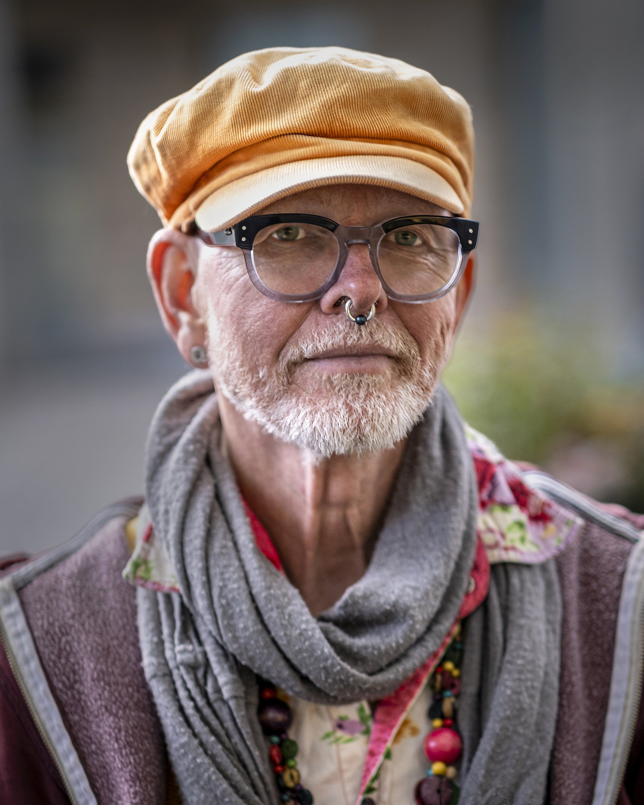 Close-up of a man with glasses, a septum piercing, a beige cap, and layered clothing, with colorful necklaces visible.