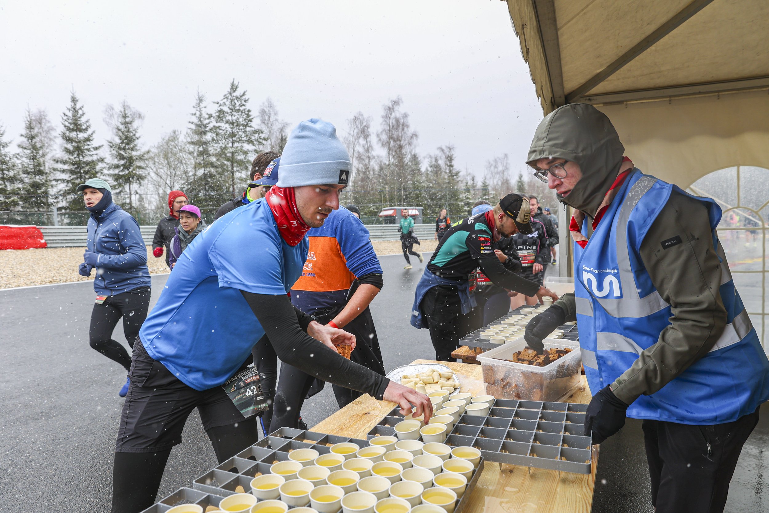 A group of people gather under a canopy at a race event on a rainy day, serving food.