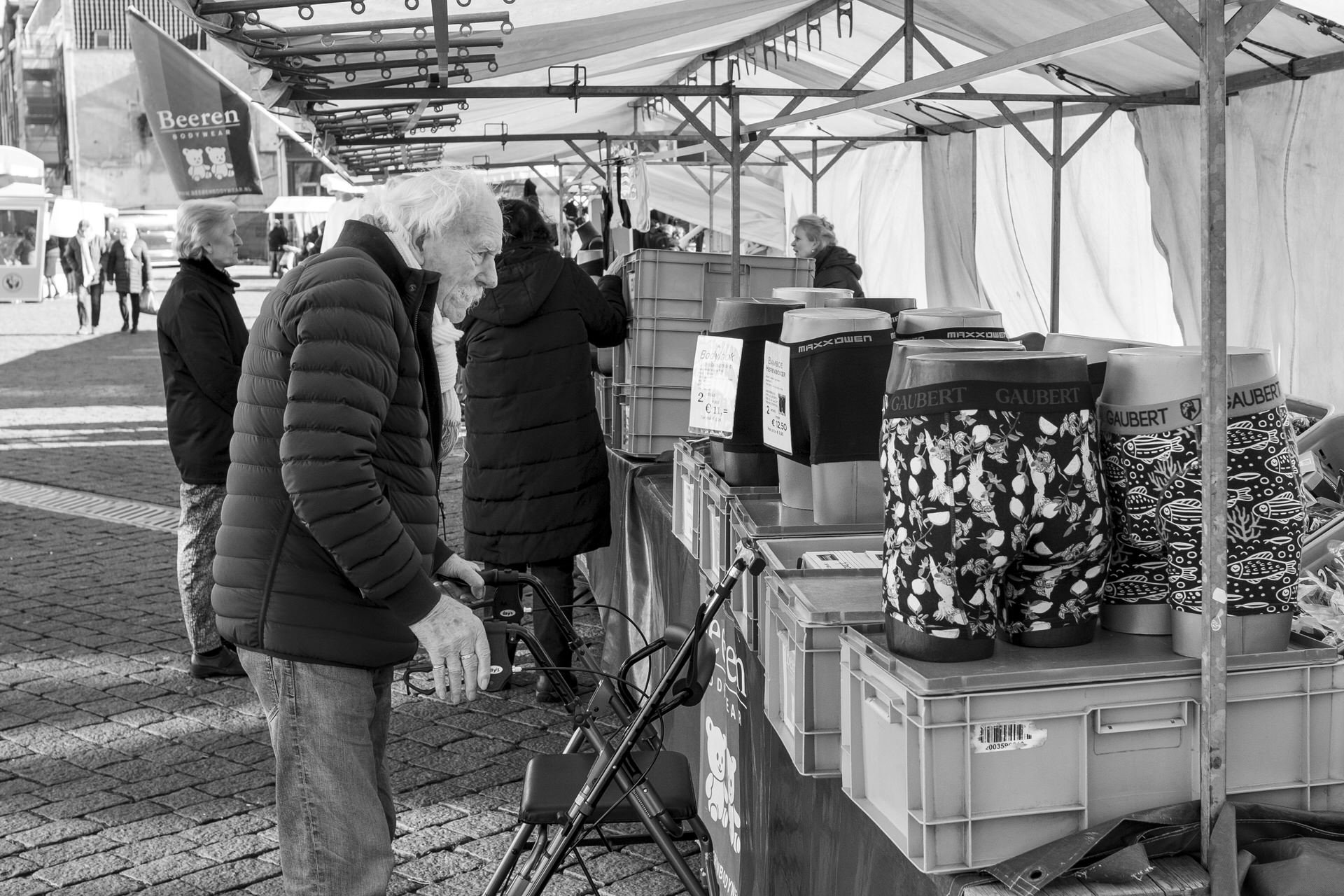 People shopping at an outdoor market stall, some with backpacks, displaying various items including socks with a fish pattern in black and white.