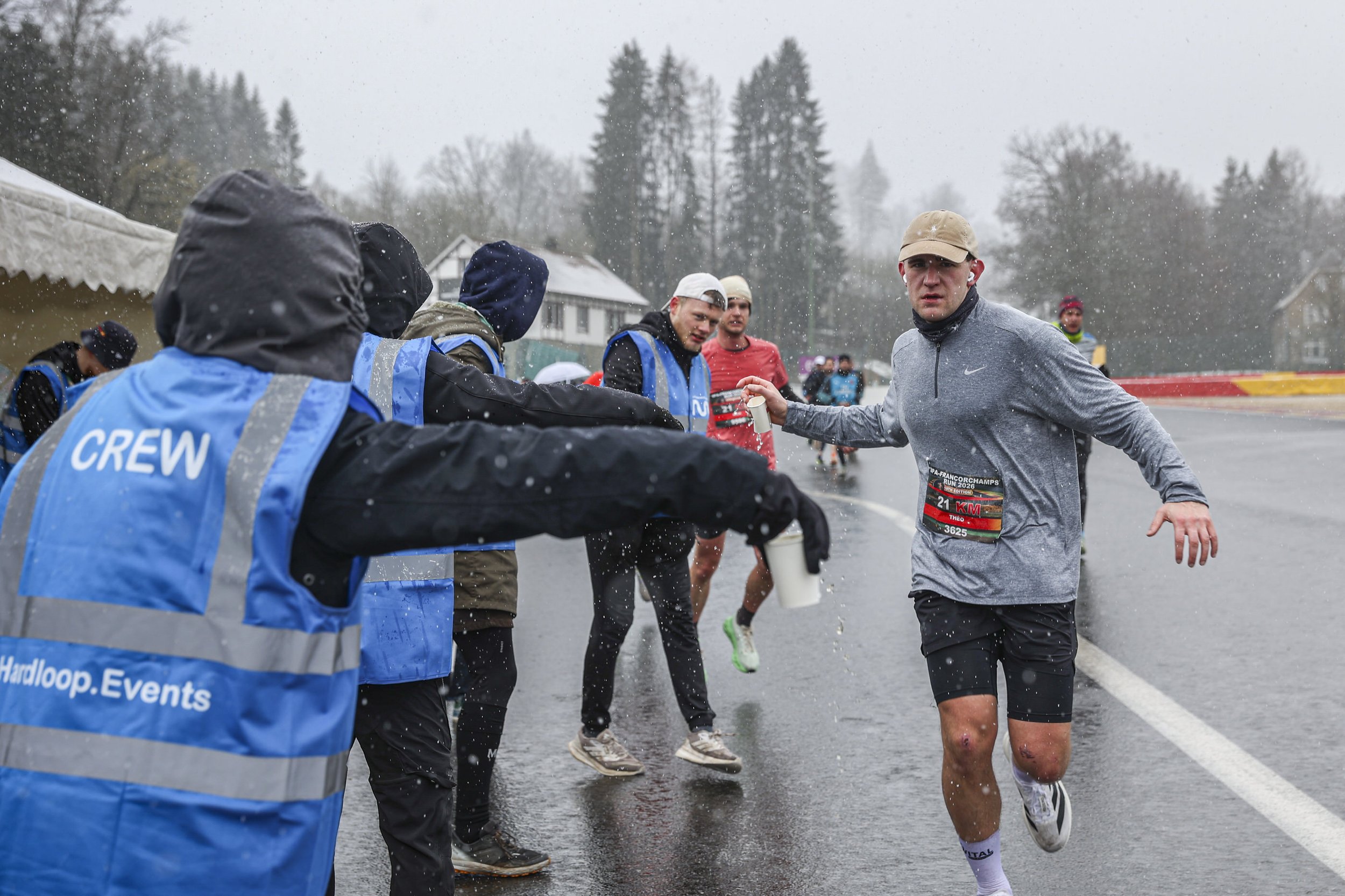 A runner in a gray Nike shirt and beige cap participating in a race in rainy weather, being handed water by volunteers in blue vests labeled 'CREW' lined up along the roadside.