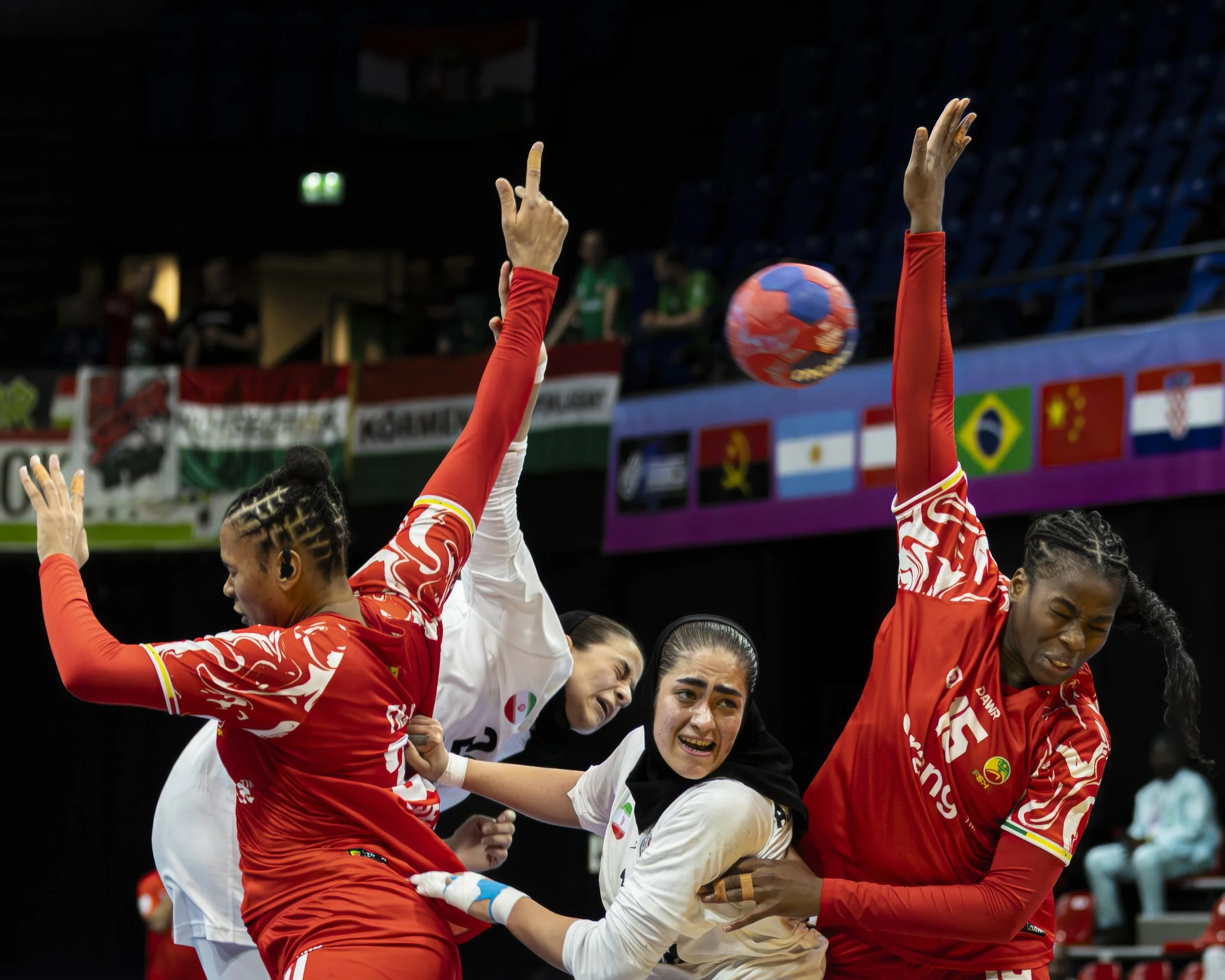 A women's volleyball team in red and white uniforms celebrating a point during a match, with a colorful volleyball in midair above them.