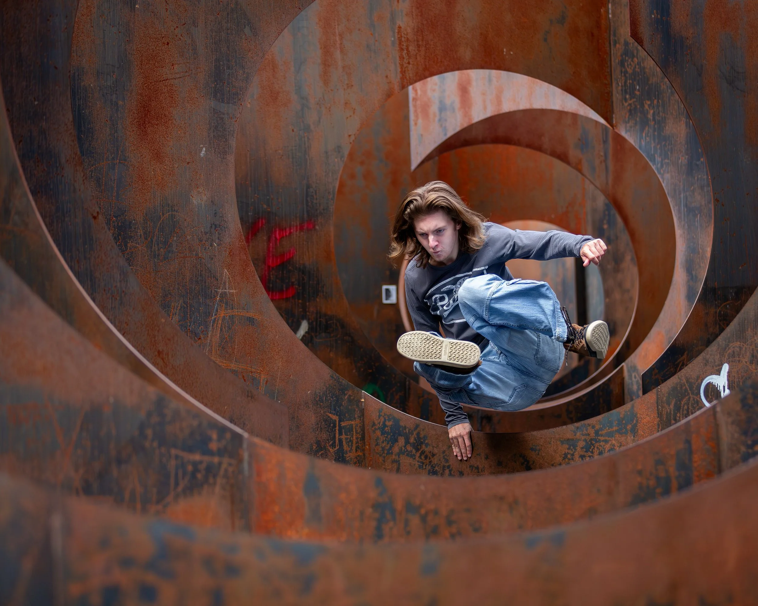 A person with long hair in a grey sweatshirt and blue jeans is performing a kick on a rusty metal sculpture with large circular openings.