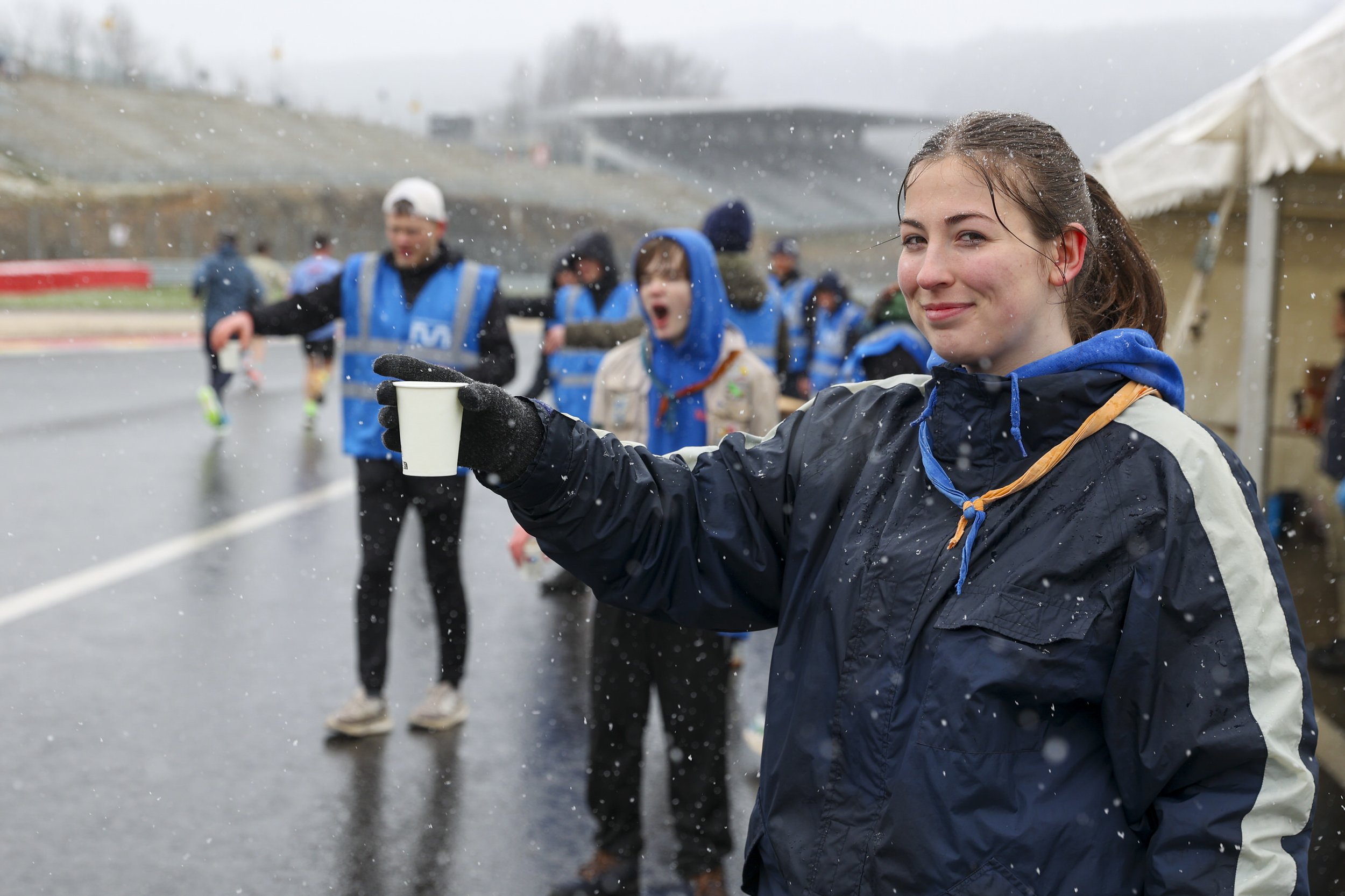 Young woman holding a cup smiling outdoors on rainy day, crowd of people in blue jackets in background at a racing event.