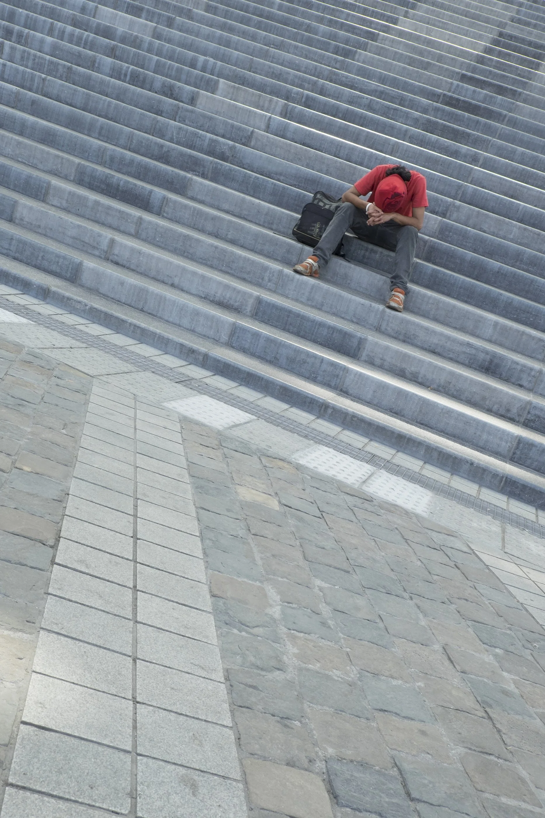 A person sitting on stairs, wearing a red shirt and red cap, with a bag beside them, head bowed and hands clasped.
