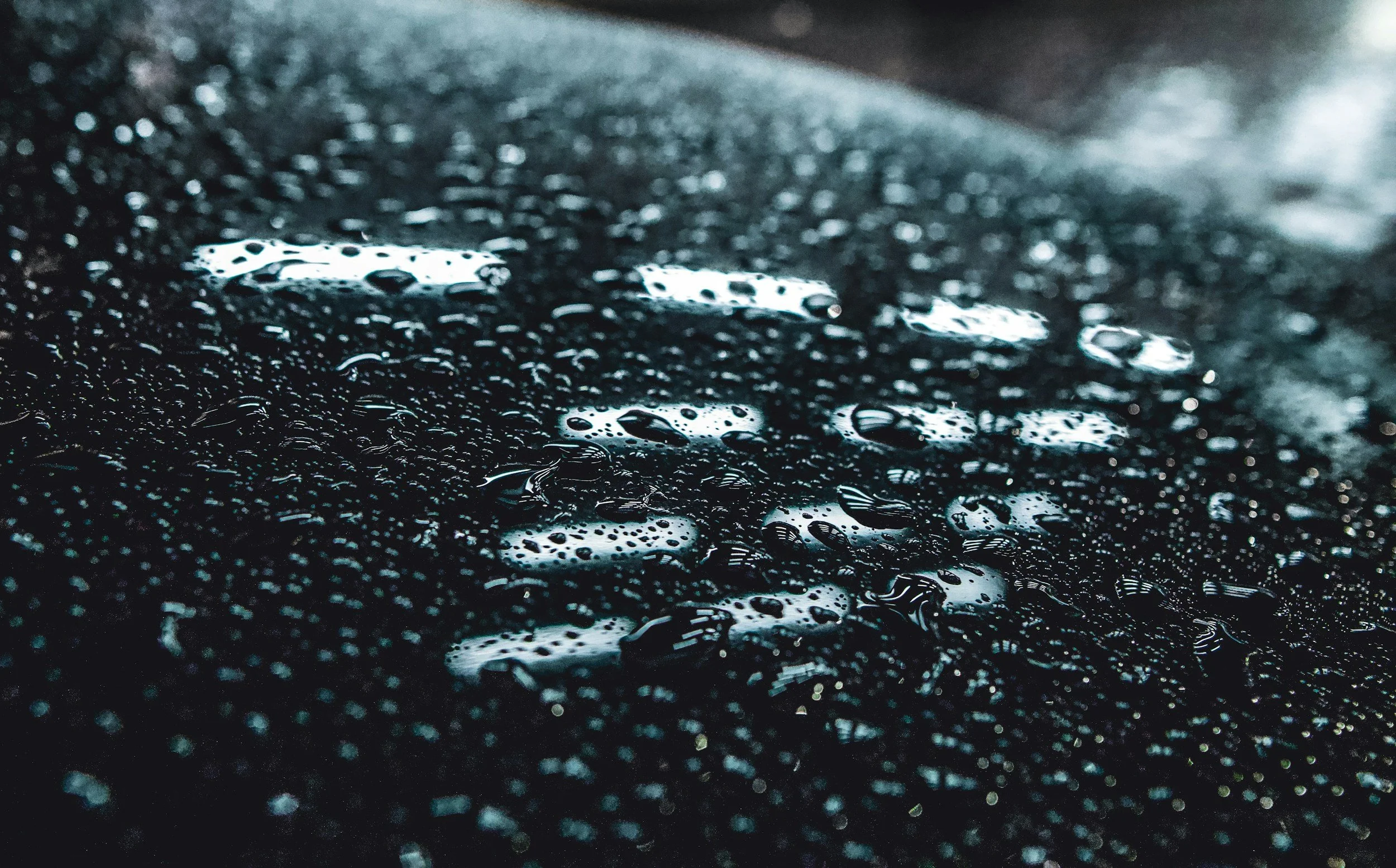 Close-up view of water droplets on a dark surface, with some droplets reflecting light, creating a textured and shiny appearance.