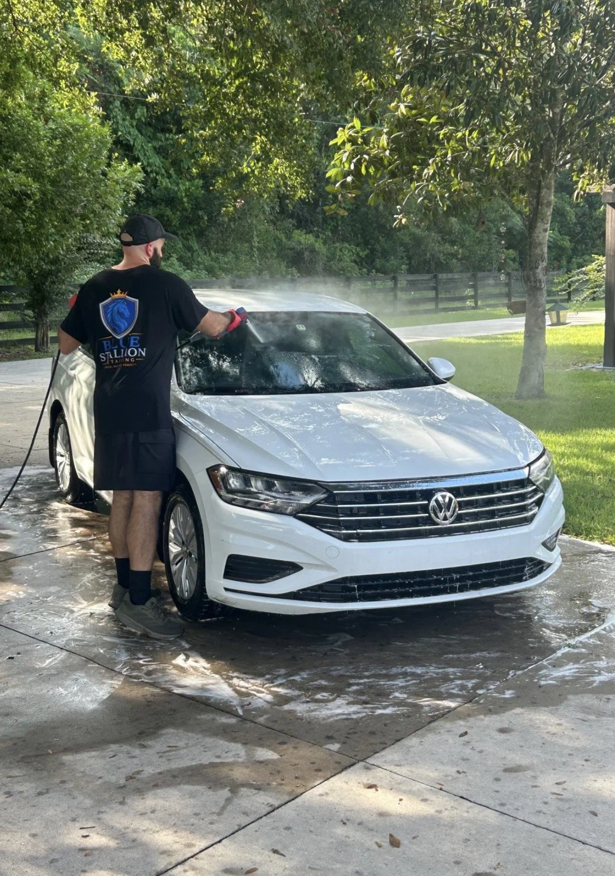A man washing a white Volkswagen sedan in a driveway surrounded by trees and greenery.