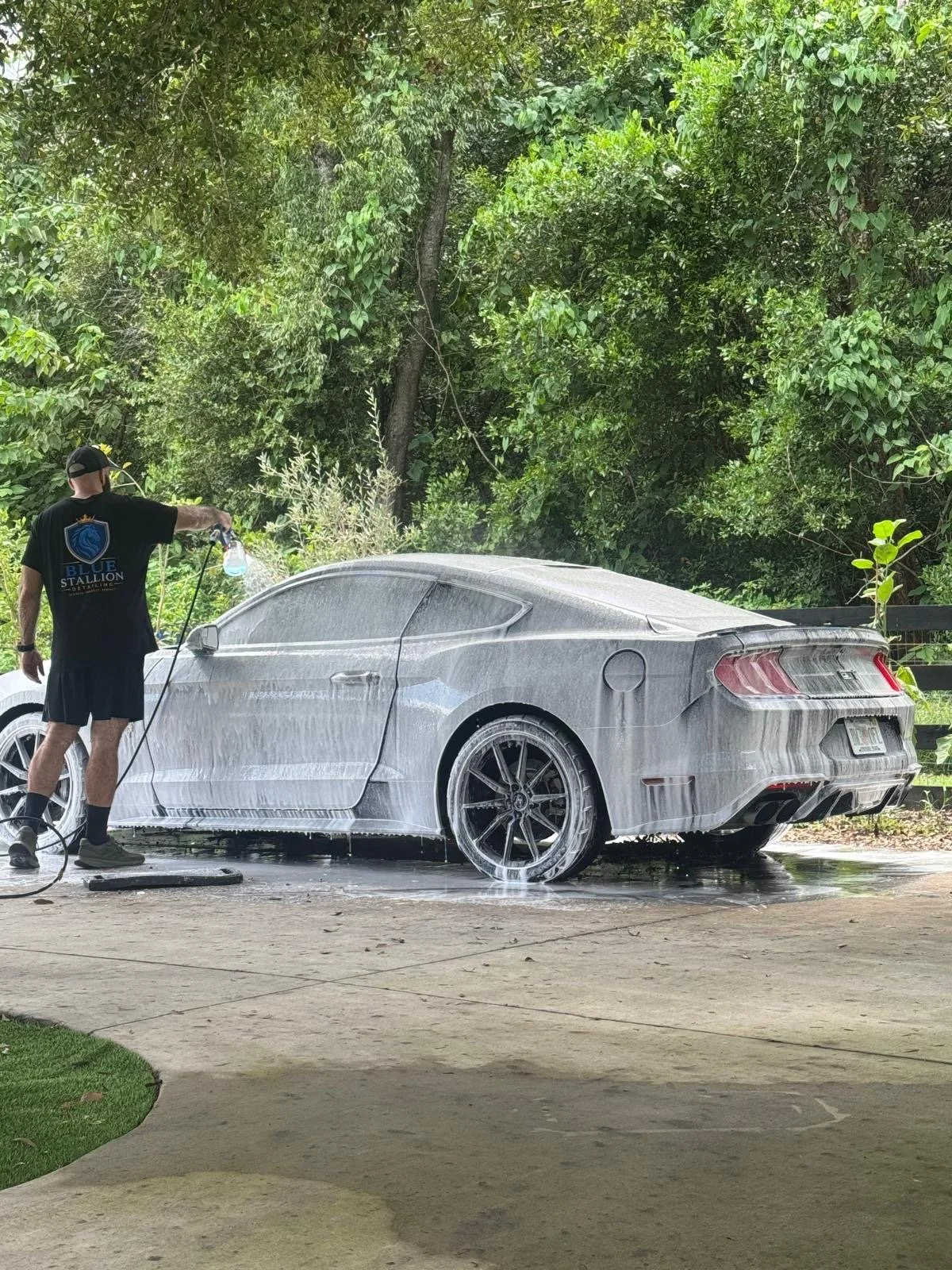 A person washing a gray sports car with foam and water outdoors next to a wooded area.