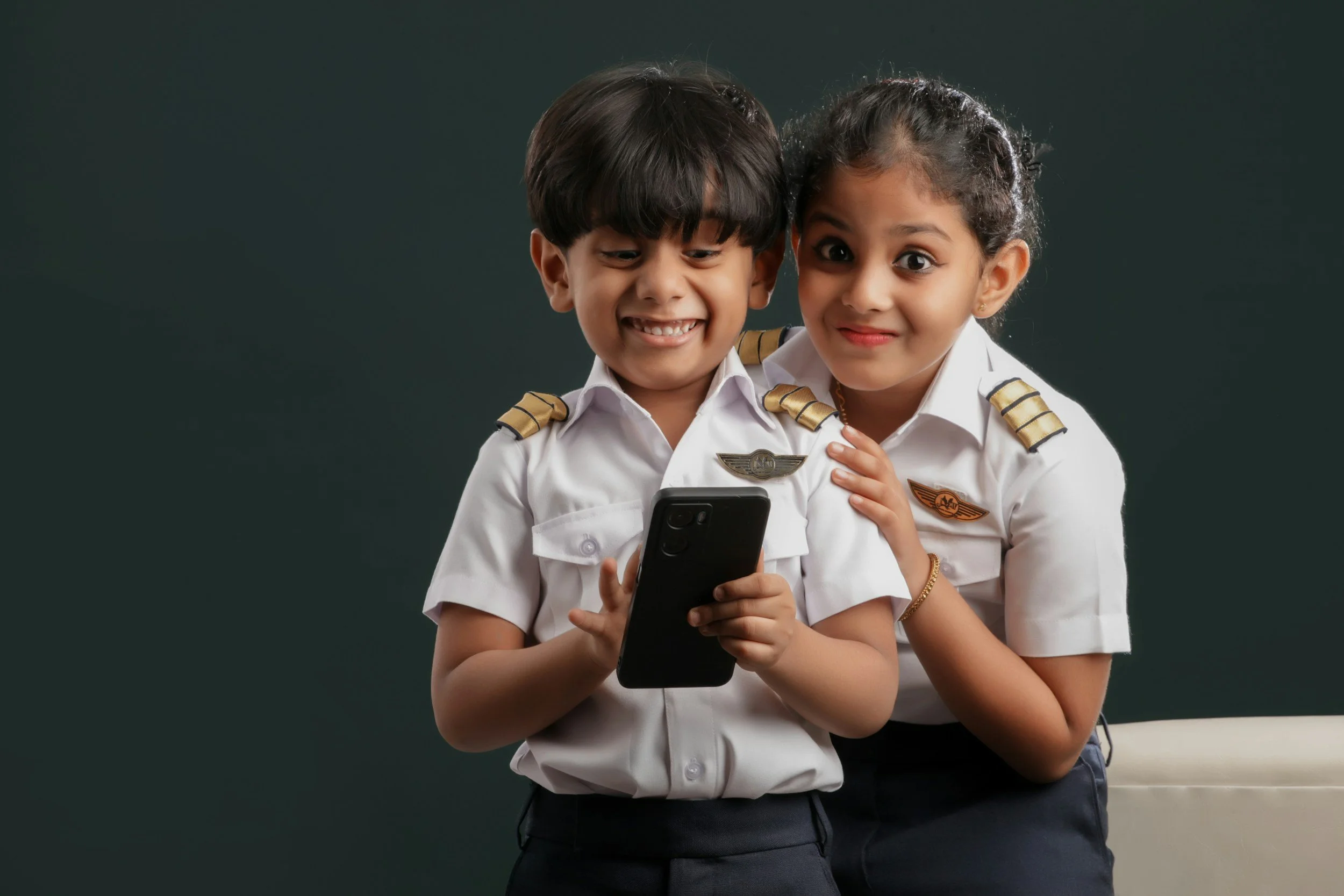 Two airline pilots, a young boy and a young girl in pilot uniforms, looking at a smartphone together while standing in front of a dark solid background.