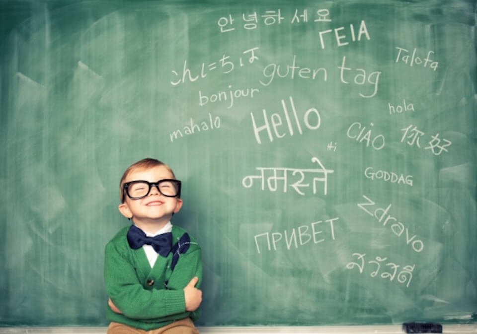 A young boy in glasses, green sweater, and bow tie, smiling in front of a green chalkboard with the word 'Hello' and the word 'Hello' in various languages written on it.