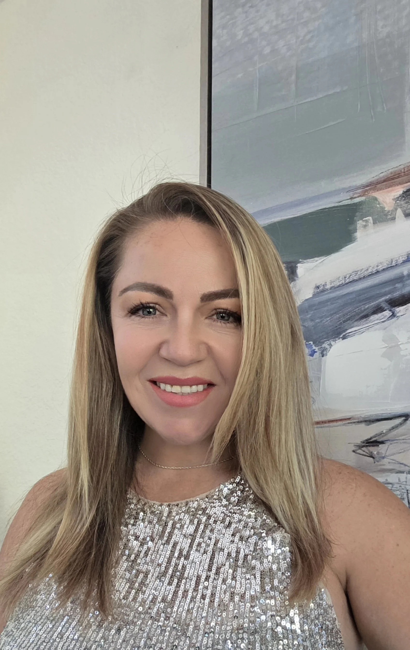 A woman with long blonde hair, smiling, wearing a sparkly silver top and a thin necklace, standing in front of a wall with abstract art.