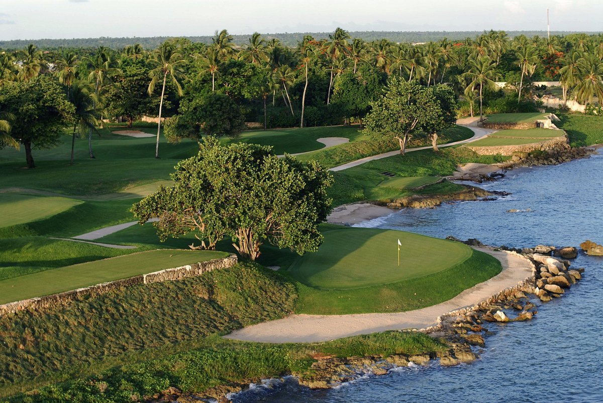 A scenic view of a golf course along a coastal shoreline with green fairways, sand traps, and a flag on the green next to the water, surrounded by numerous trees and palm trees in the background.