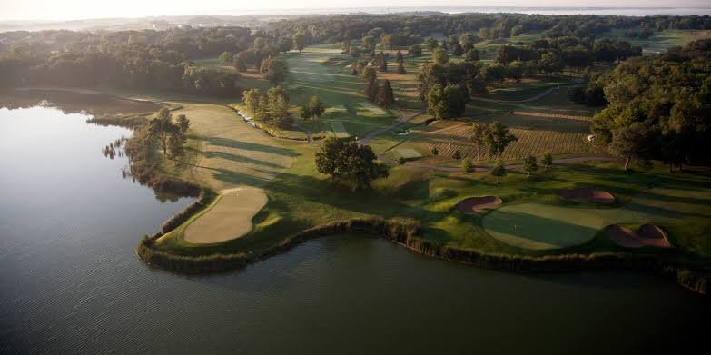 Aerial view of a golf course next to a lake, with lush green fairways, sand bunkers, trees, and surrounding farmland in the distance.