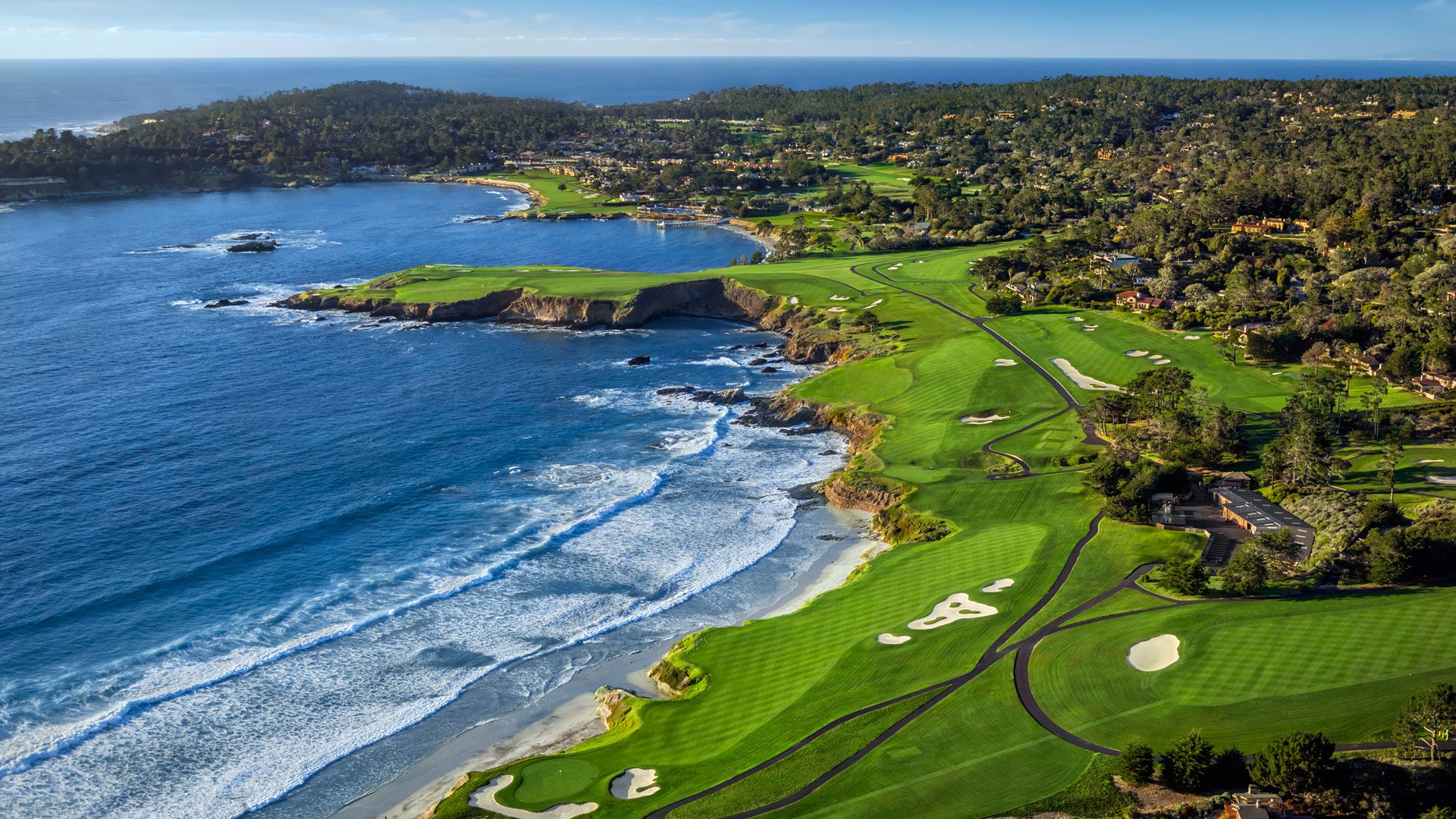 Aerial view of a coastal golf course with green fairways and white sand bunkers along the rocky shoreline, adjacent to the ocean with waves crashing onto the beach, trees, and residential neighborhoods in the background.
