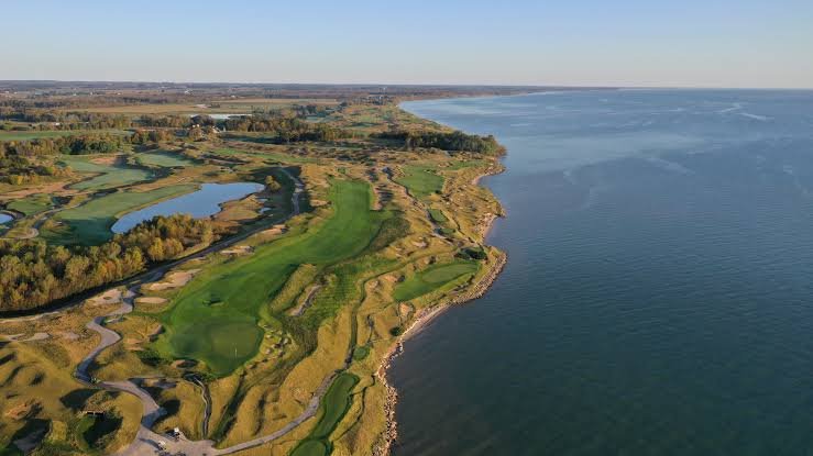 Aerial view of a golf course along a coastline, with water on one side and lush green fairways and sand traps on the other.