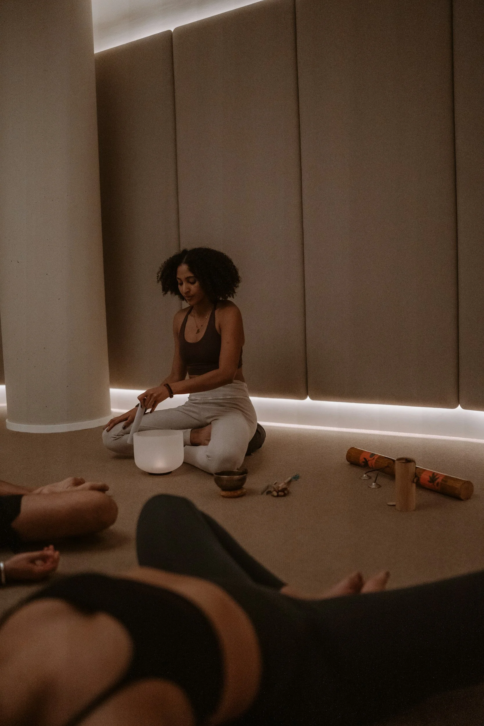 A woman with curly hair is practicing meditation or sound healing in a dimly lit room, sitting cross-legged on the floor with a white crystal singing bowl in front of her. Items such as rolled-up yoga mats, bowls, and small objects are scattered around her.