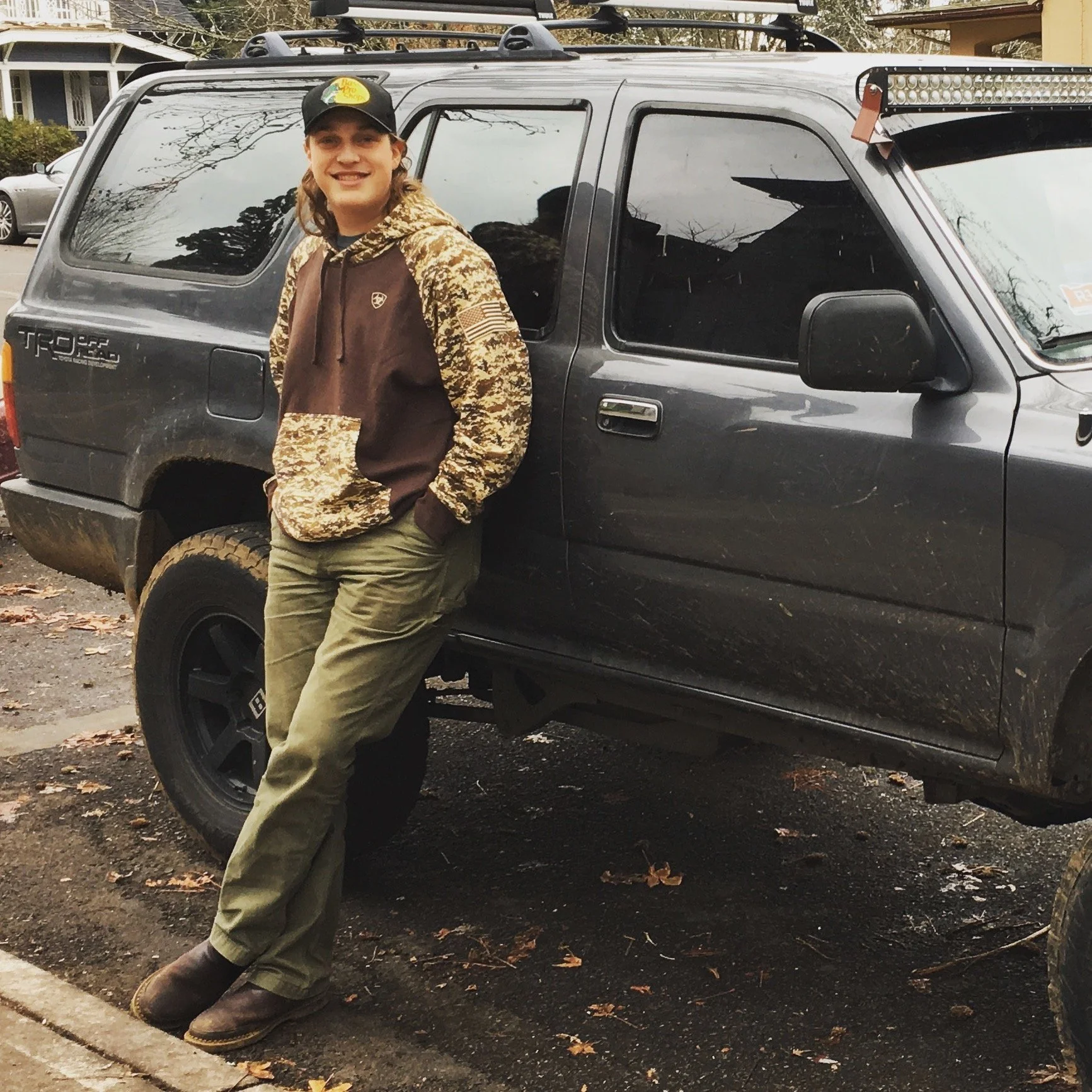 A smiling young man with long hair wearing a hat and camouflage-patterned hoodie, standing outdoors with his hands in his pockets, leaning against a black Toyota T100 pick-up truck in a suburban neighborhood with fallen leaves on the ground.