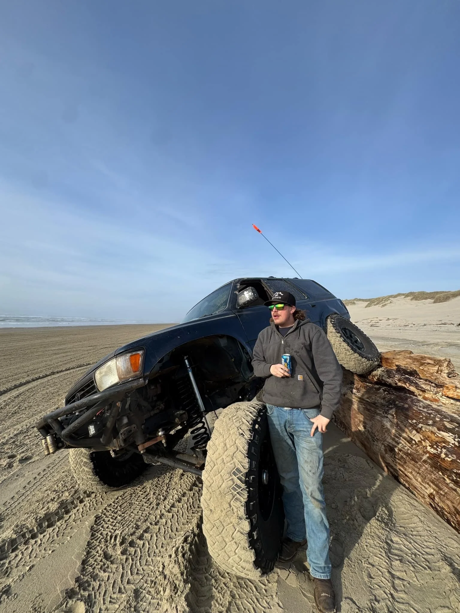 Person in a gray hoodie and blue jeans standing next to a black off-road vehicle that is overturned on a sandy beach, with a large fallen log behind the vehicle. The person is holding a drink can and wearing sunglasses and a baseball cap. The sky is 