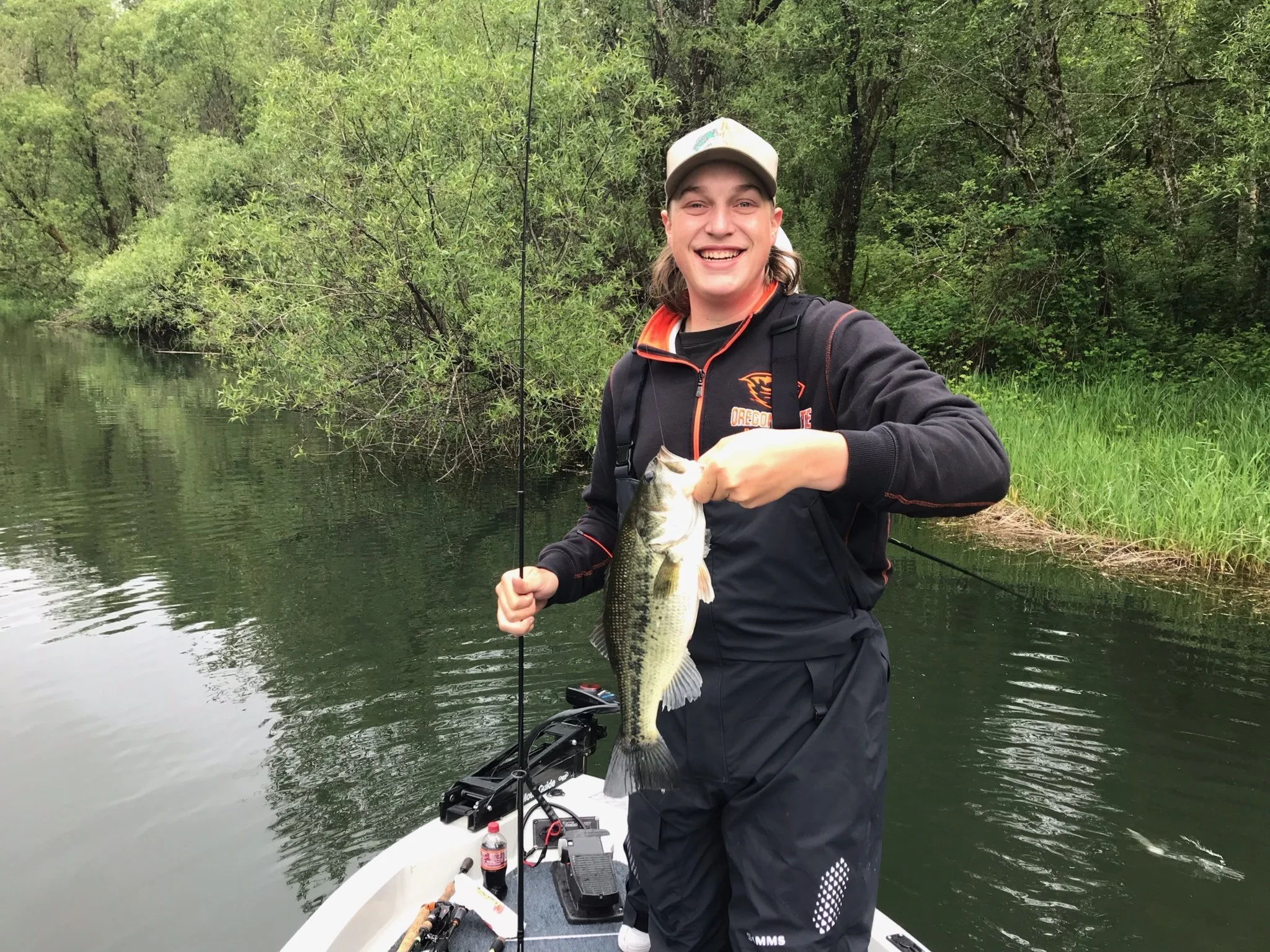 A young man smiling and holding a large fish while fishing on a small boat in a river with dense green trees in the background.