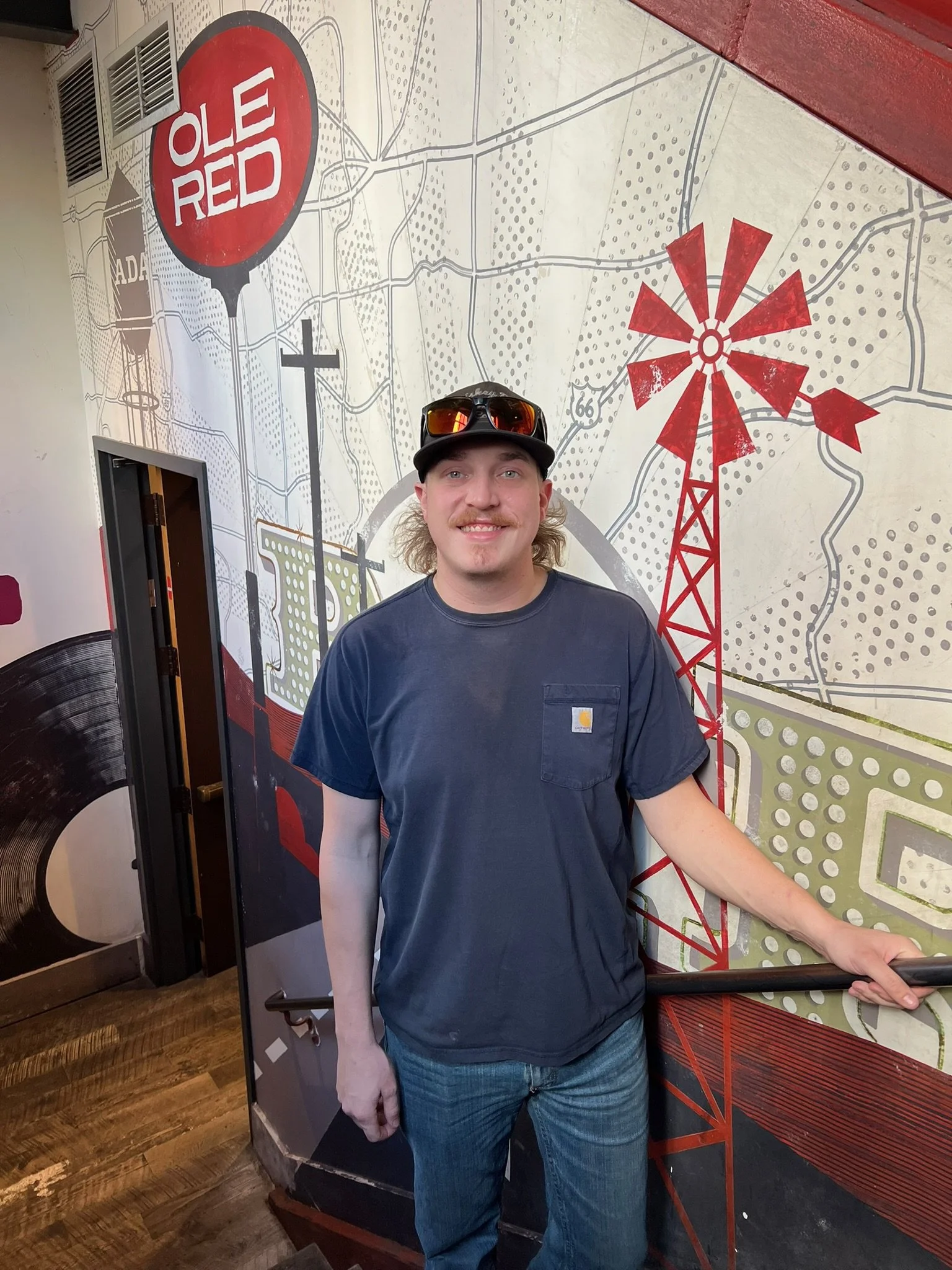 A man wearing a black cap with orange sunglasses on top, a dark blue Carhartt t-shirt, and jeans, standing indoors in front of a wall with a mural of a map and the words "Old Red" inside a red speech bubble.