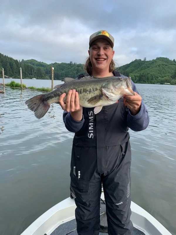 A person holding a large fish on a boat, on a lake with green hills and trees in the background under cloudy skies.