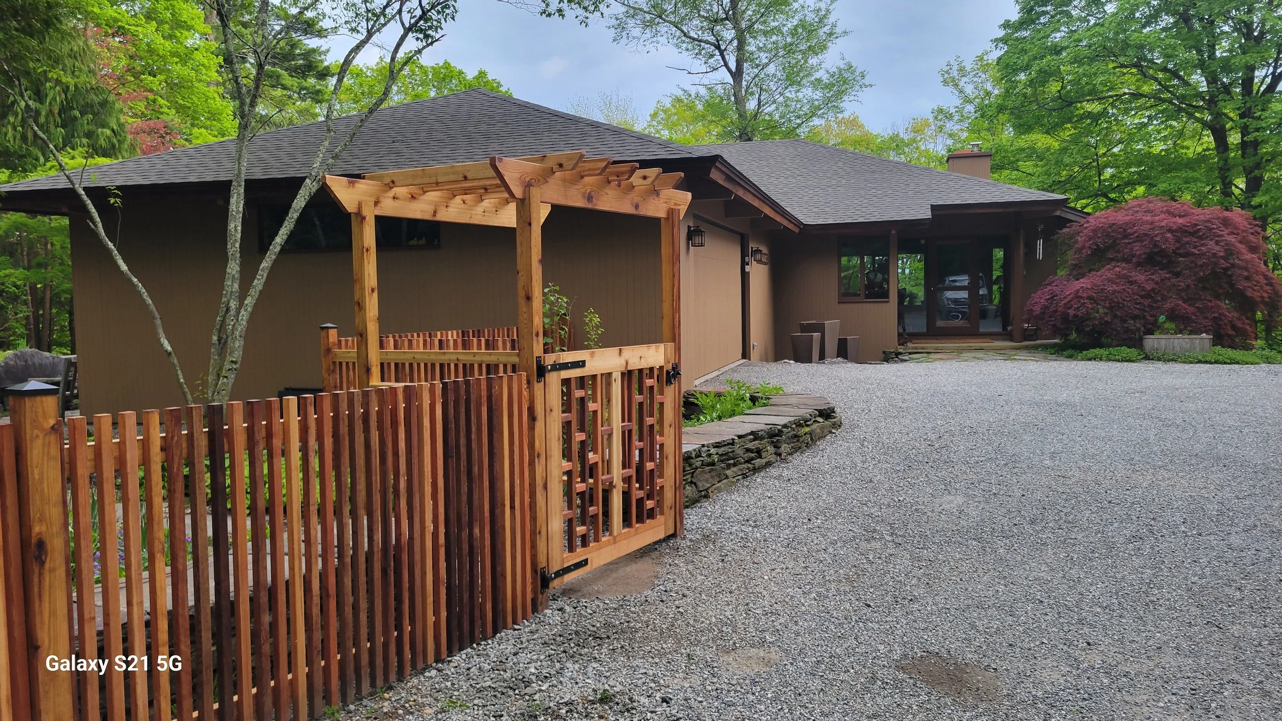 A house with a gravel driveway, surrounded by lush green trees and bushes, with a partially constructed wooden pergola in the front yard.