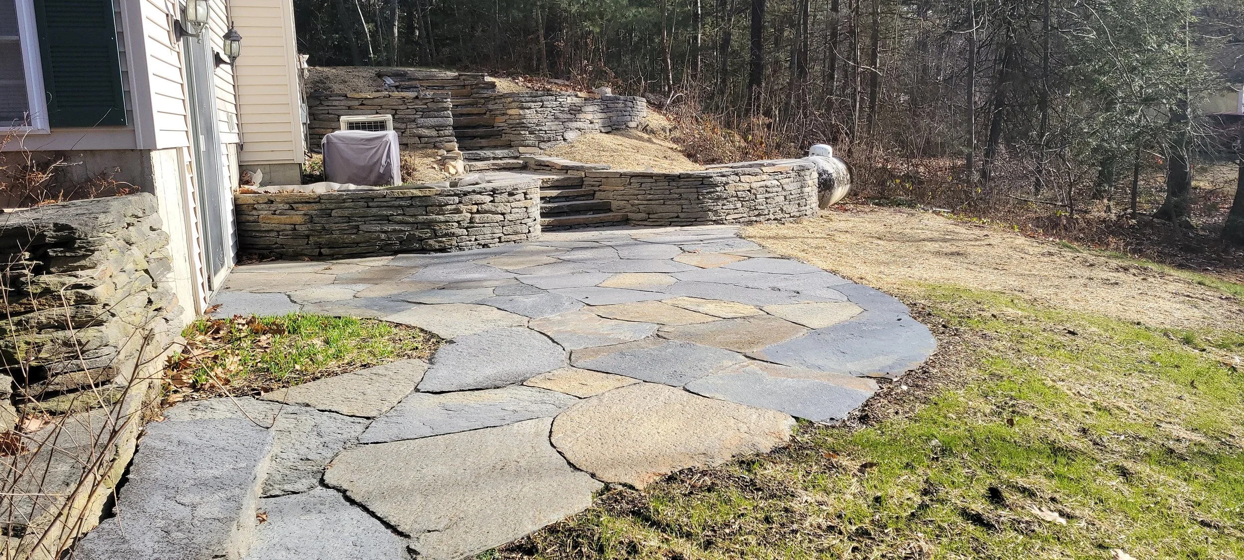 Stone patio with steps leading up to a landscaped backyard with stone retaining walls and surrounding trees.