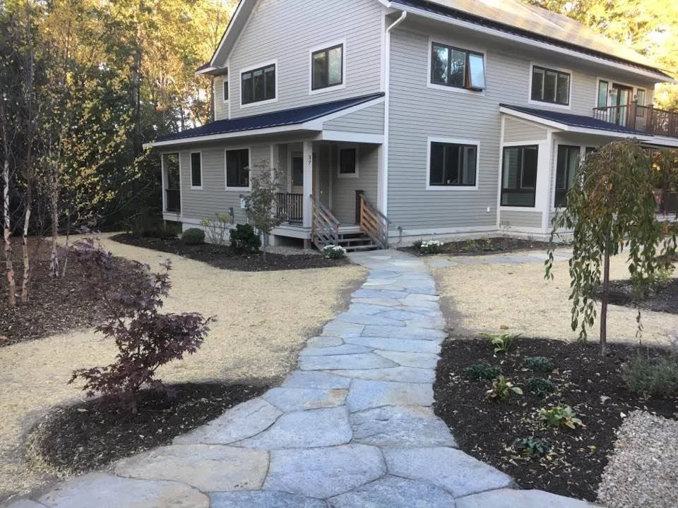 A front yard with a stone pathway leading to a two-story house with gray siding, surrounded by trees and landscaped plants.
