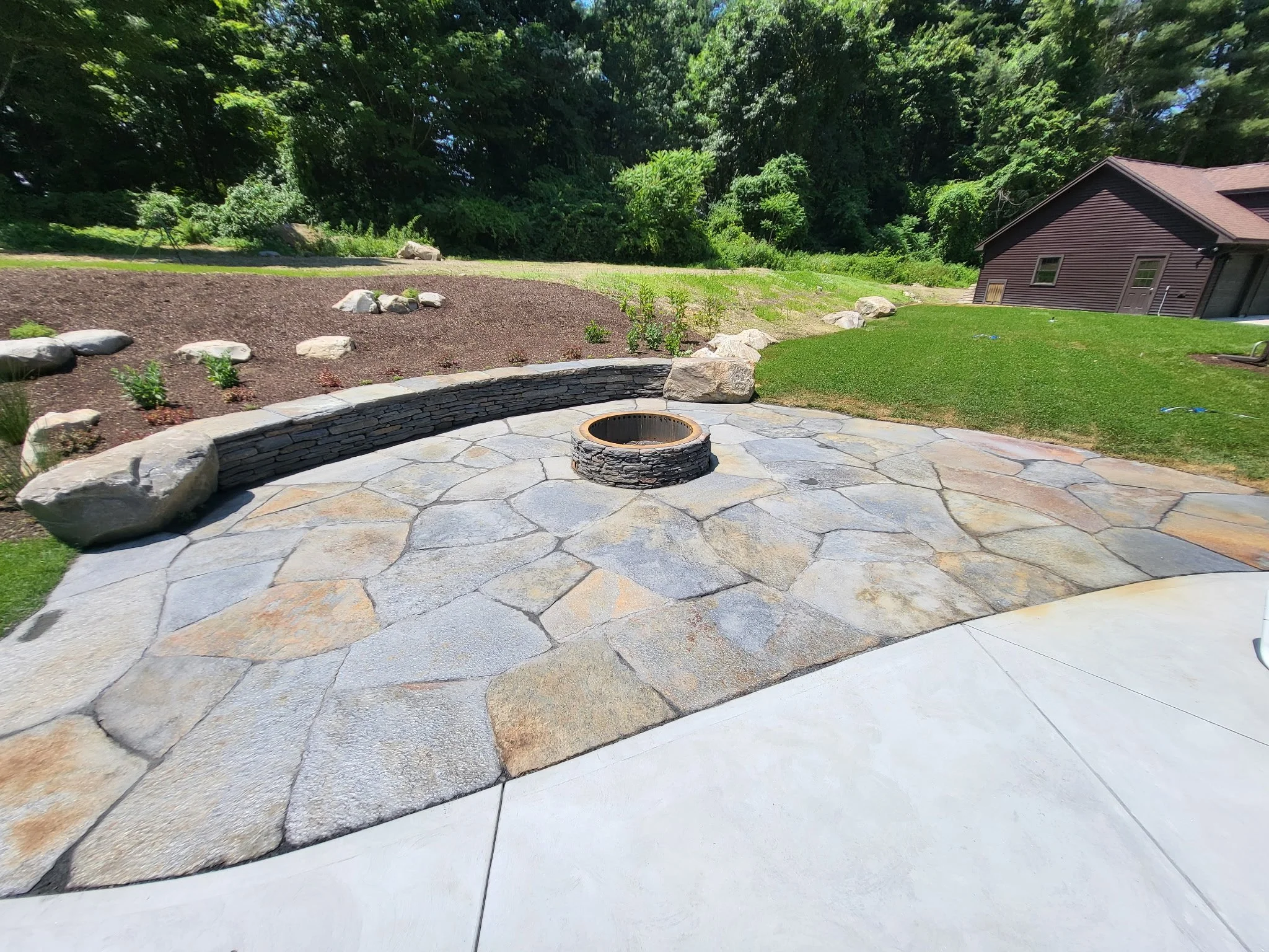 A backyard patio with a stone fire pit, a landscaped garden area with small plants and rocks, a grassy yard, a brown house with a sloped roof, and a backdrop of dense green trees.