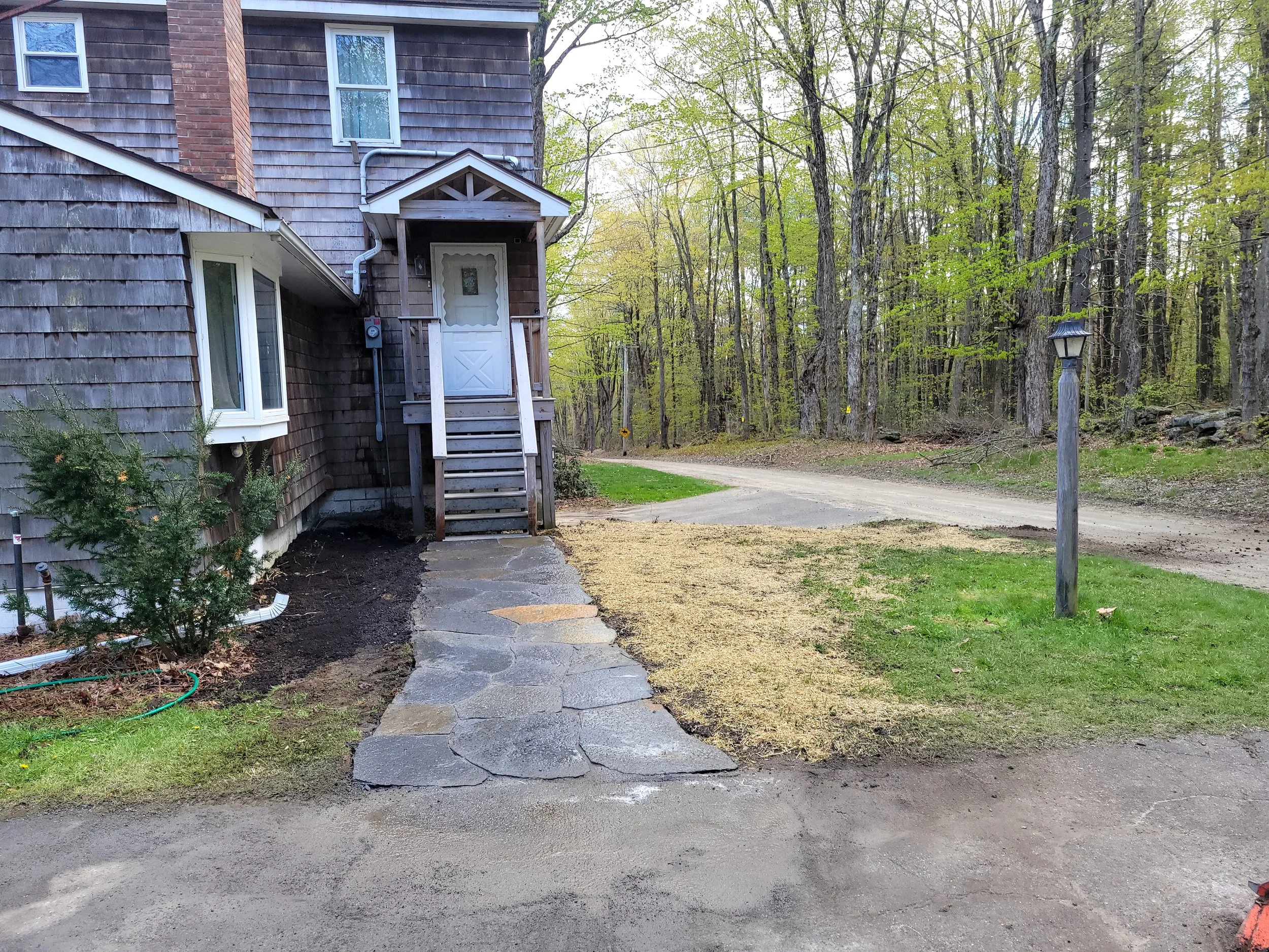 Exterior side of a house with weathered wooden siding, a small porch with stairs, and a white door. The yard has a stone pathway, a small pine tree, and a patch of dirt. There is a dirt driveway and a forest background with tall trees.