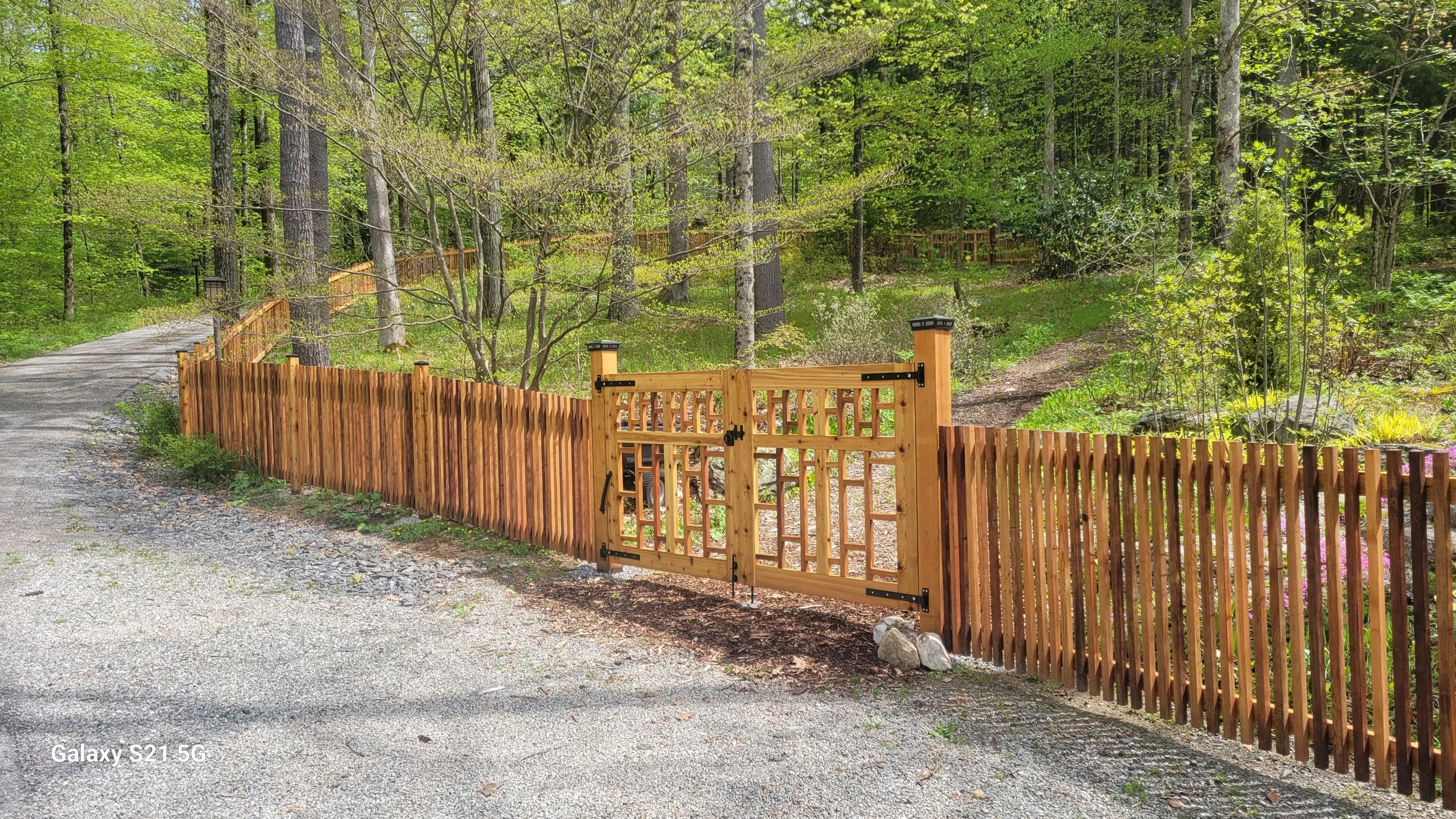 A wooden gate with latch and hinges, set in a wooden fence, opening into a lush green forest with tall trees, bushes, and a gravel driveway in the foreground.