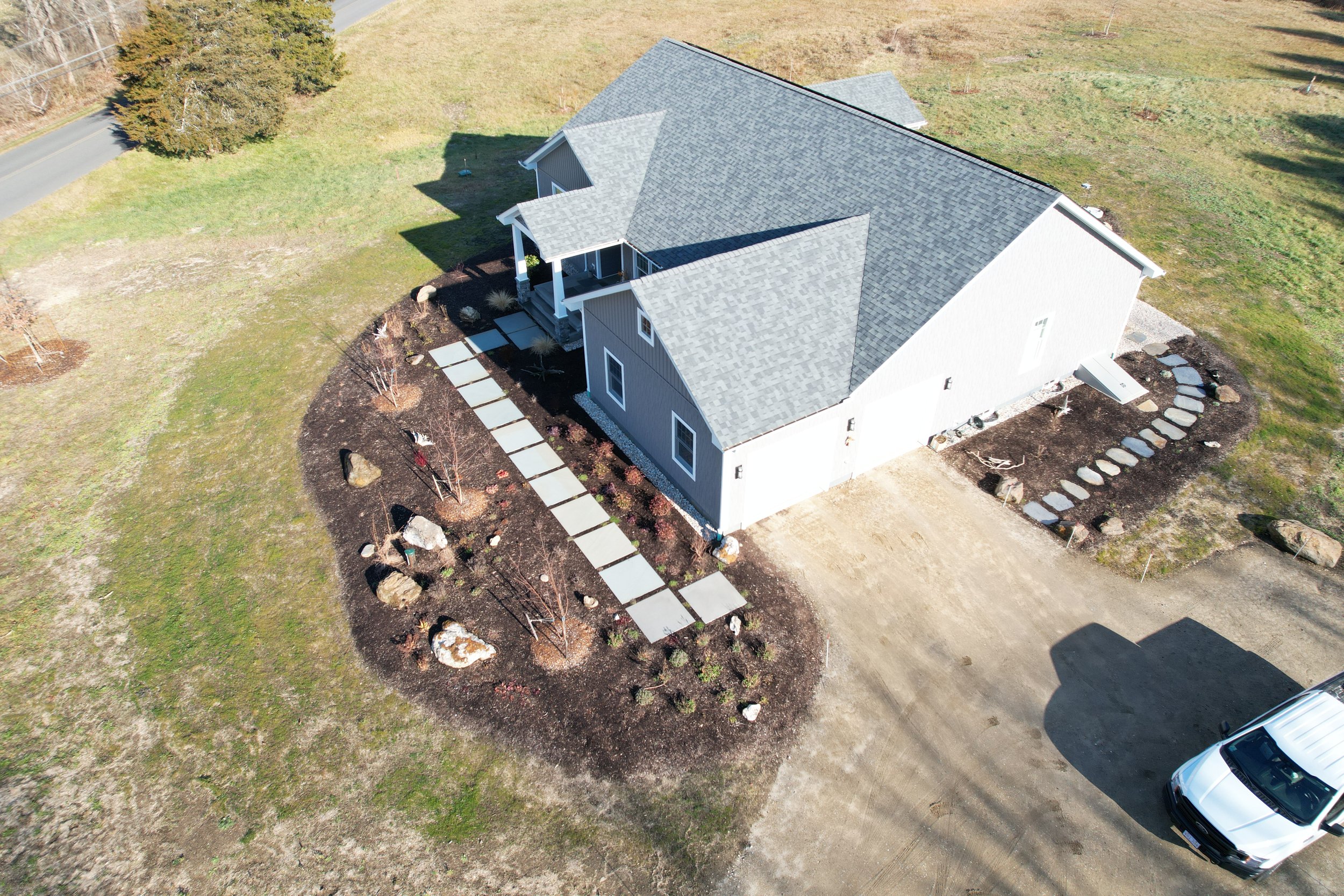 Aerial view of a house in a semi-rural area with a gray roof, white exterior walls, and a small front porch. The yard has a landscaped garden with stone pathways and a few trees, surrounded by grassy fields and a road.