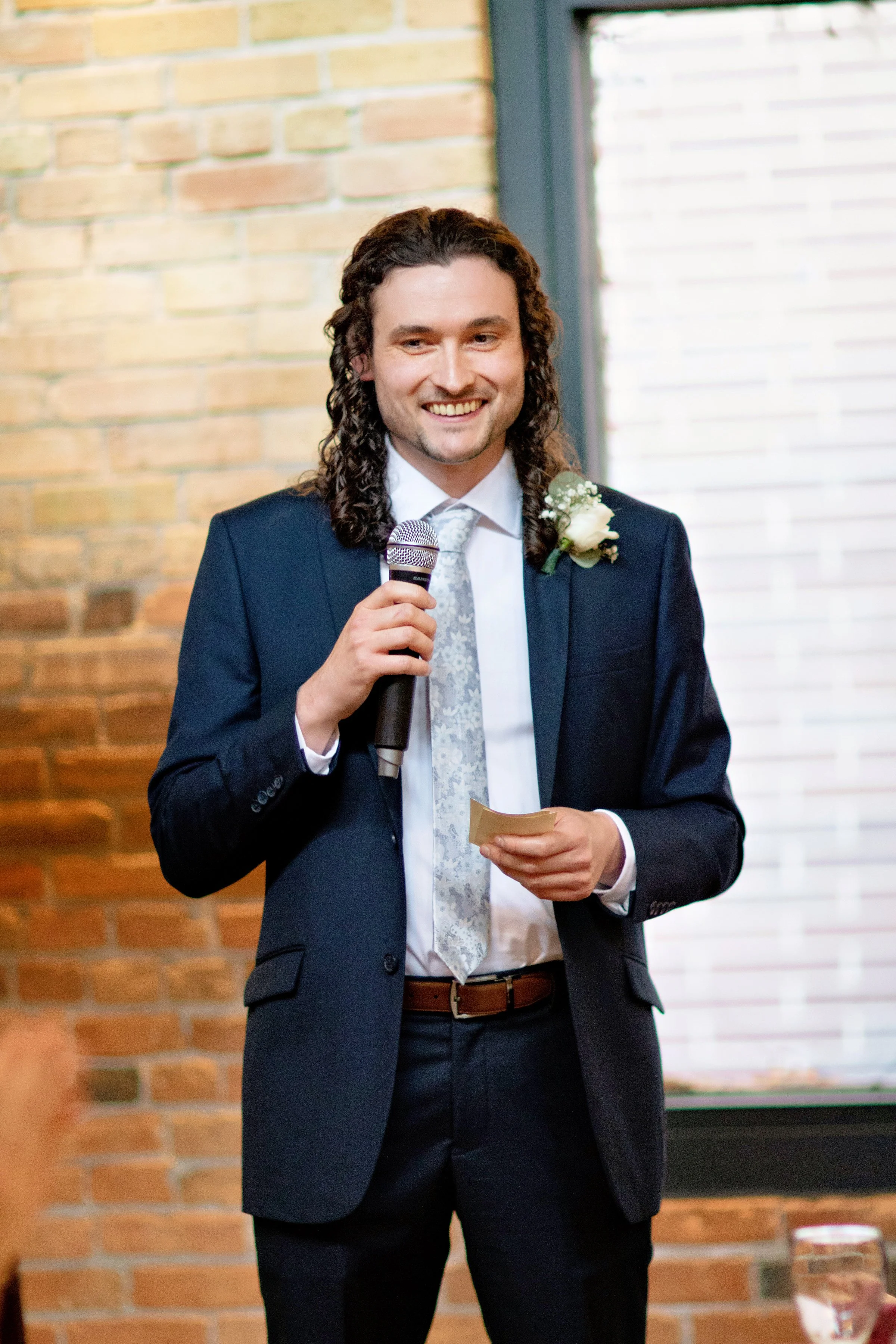 A man in a dark suit holding a microphone, smiling, with a boutonniere on his suit lapel, standing in front of a brick wall and window.