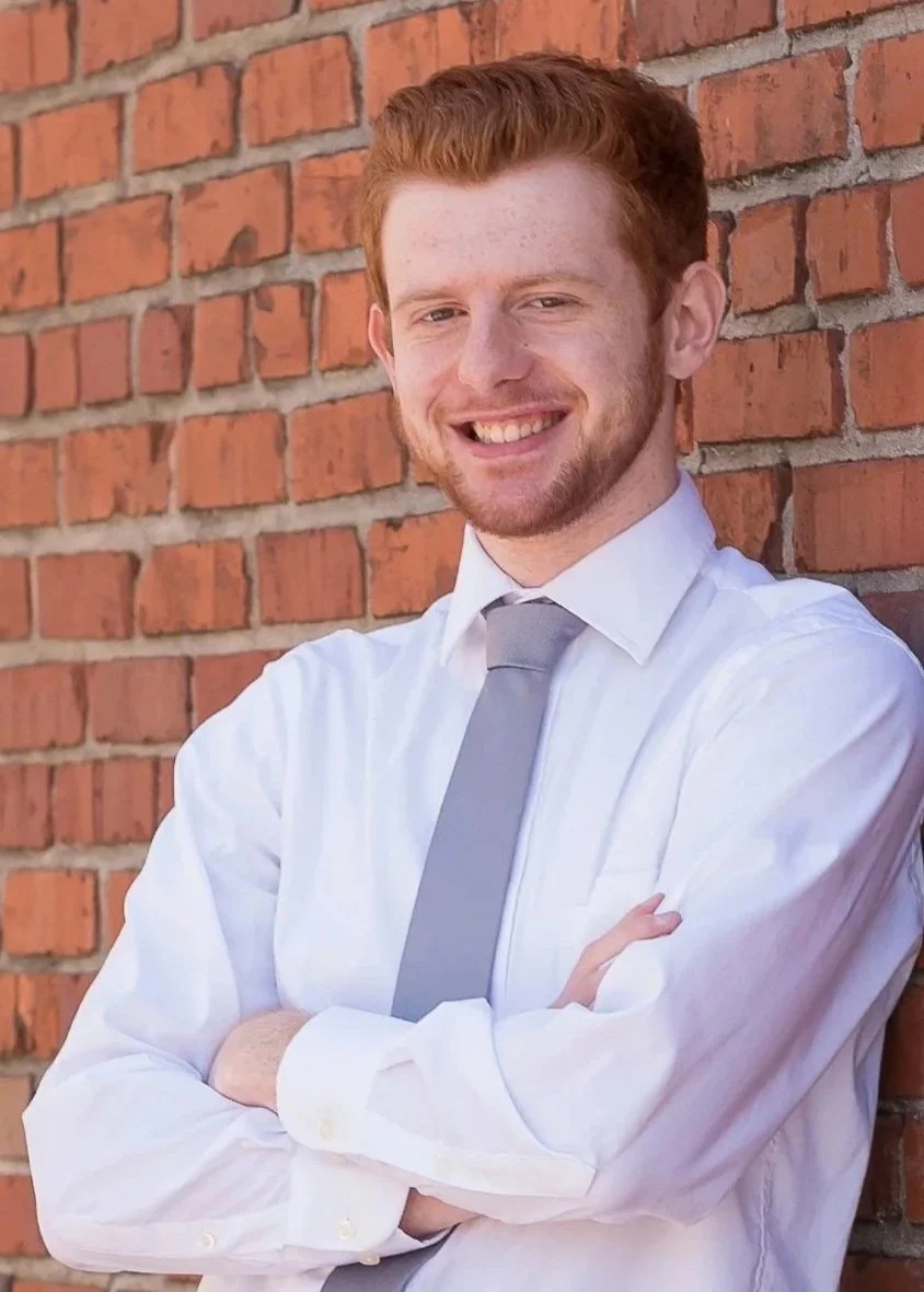 Young man with red hair and beard, smiling, standing against a brick wall, wearing a white shirt and gray tie.