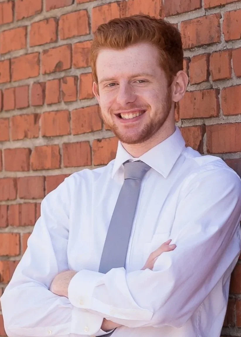 Young man with red hair, beard, and a white shirt, wearing a light gray tie, standing with arms crossed in front of a brick wall, smiling at the camera.
