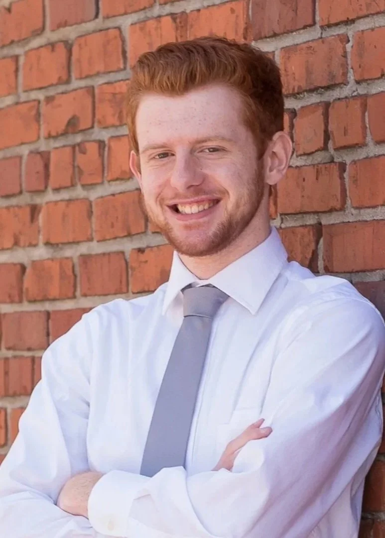 A young man with red hair and a beard smiling, wearing a white dress shirt and gray tie, standing against a red brick wall with arms crossed.