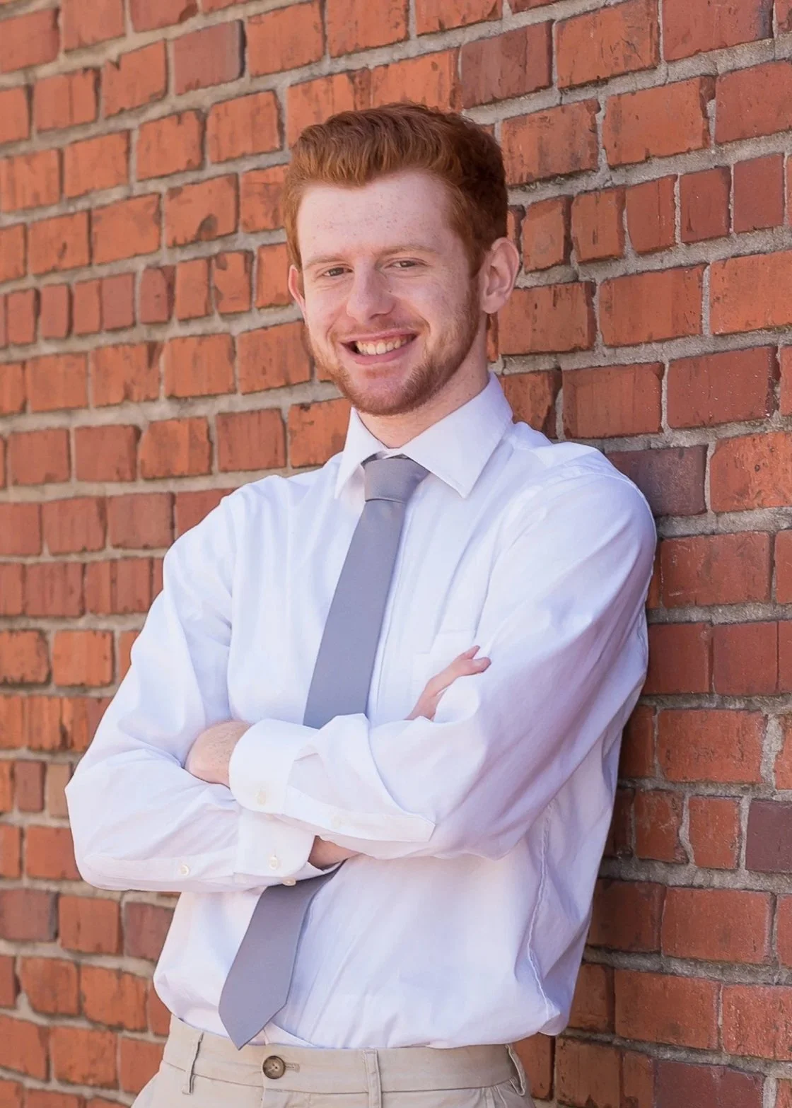 A young man with red hair and a beard standing against a brick wall, smiling, wearing a white shirt, light gray tie, and beige pants with arms crossed.