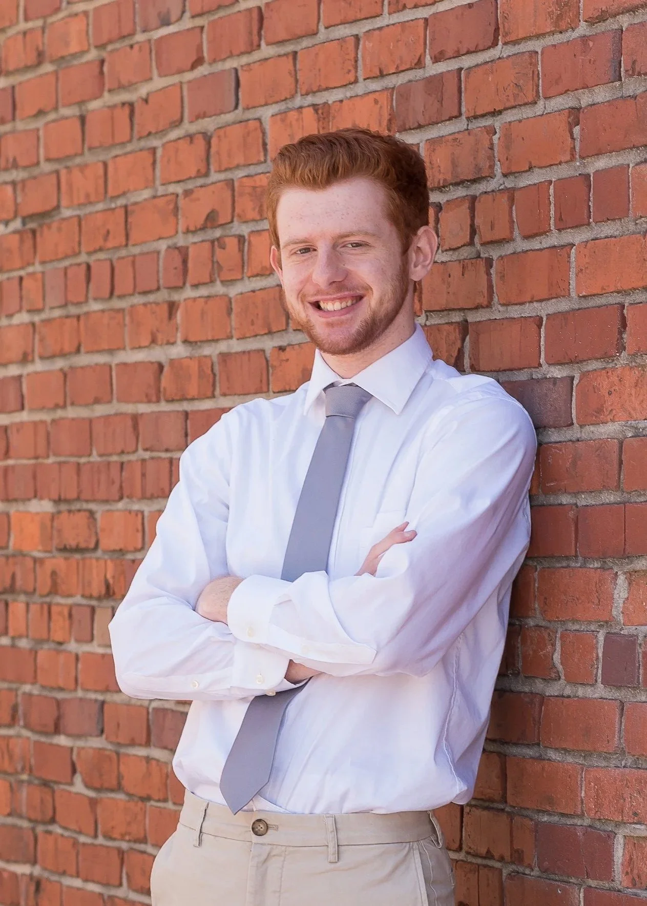 A young man with red hair and a beard, wearing a white dress shirt and a light gray tie, standing with arms crossed in front of a red brick wall, smiling.