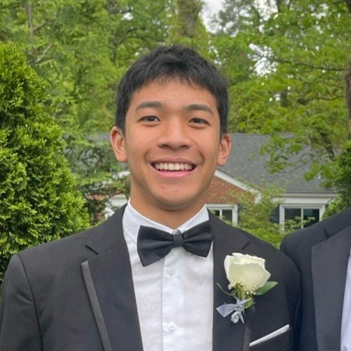 Teenage boy with short black hair, wearing a black tuxedo with a bow tie and a white shirt, smiling outdoors in a green, leafy setting, with a building in the background.