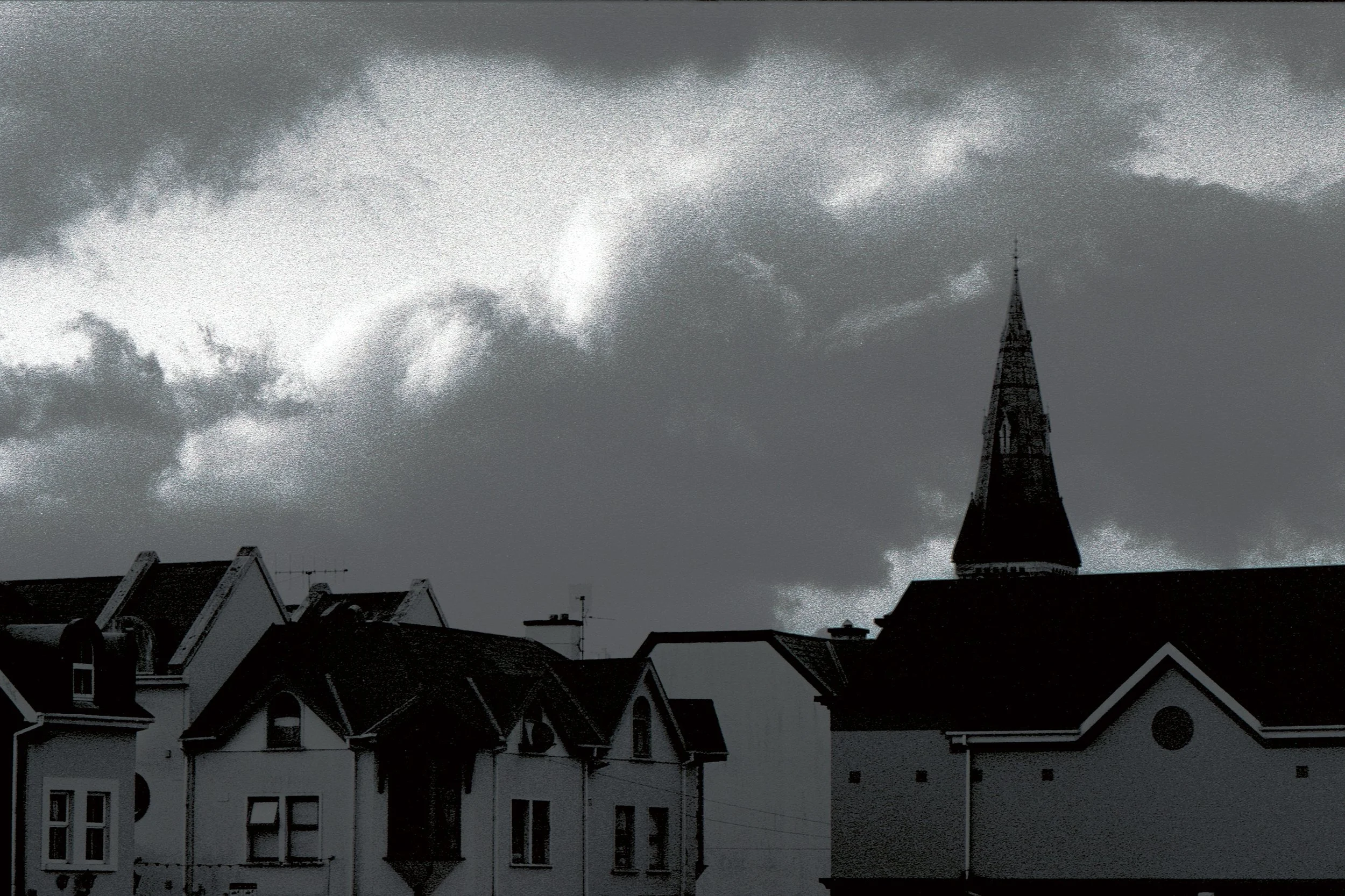 A black and white photo of a cityscape with rooftops and a church steeple under a cloudy sky.