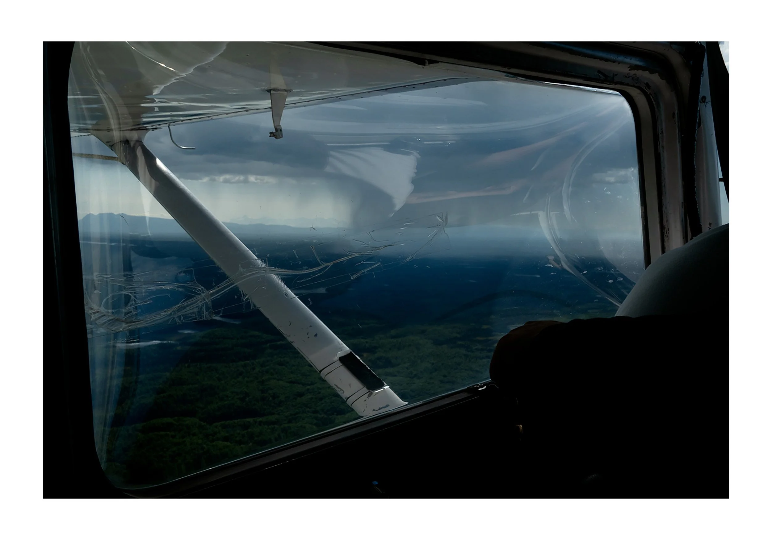 View from inside a small aircraft cockpit showing a cracked windshield and wing structure, with a landscape of green land and distant mountains below, under a cloudy sky.