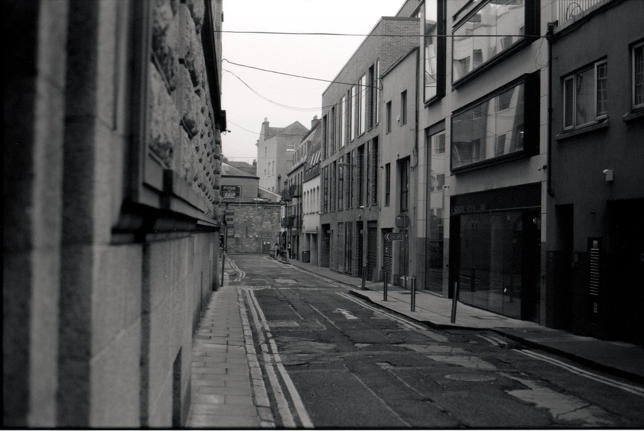 A narrow city street with modern buildings on one side and an older stone wall on the other. The street has double yellow lines and a small sidewalk, with a few street signs visible in the distance. The sky is overcast.