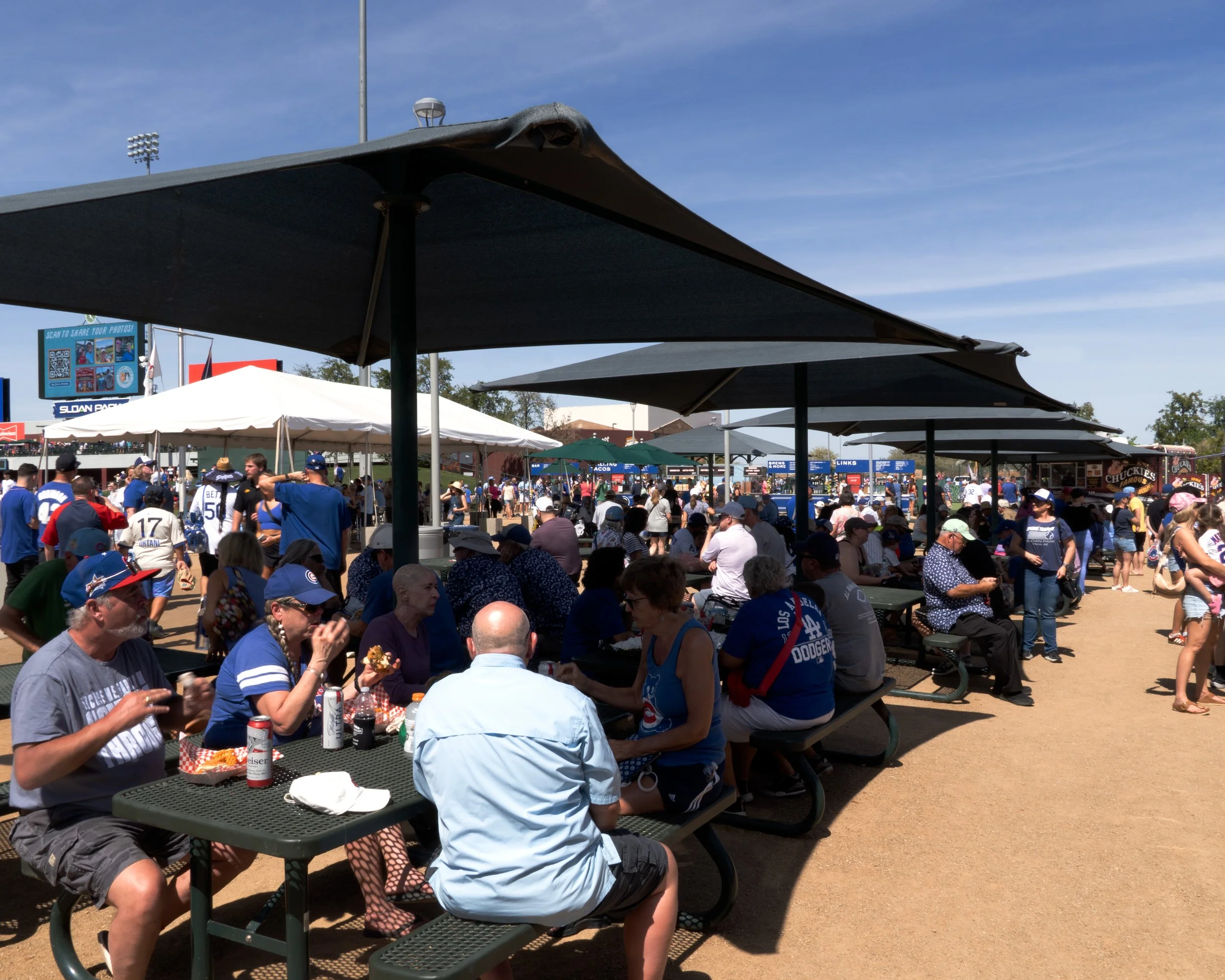 Crowd of people sitting and standing at picnic tables under large black umbrellas at an outdoor event, sunny weather with a blue sky, sports stadium lights, and signs in the background.