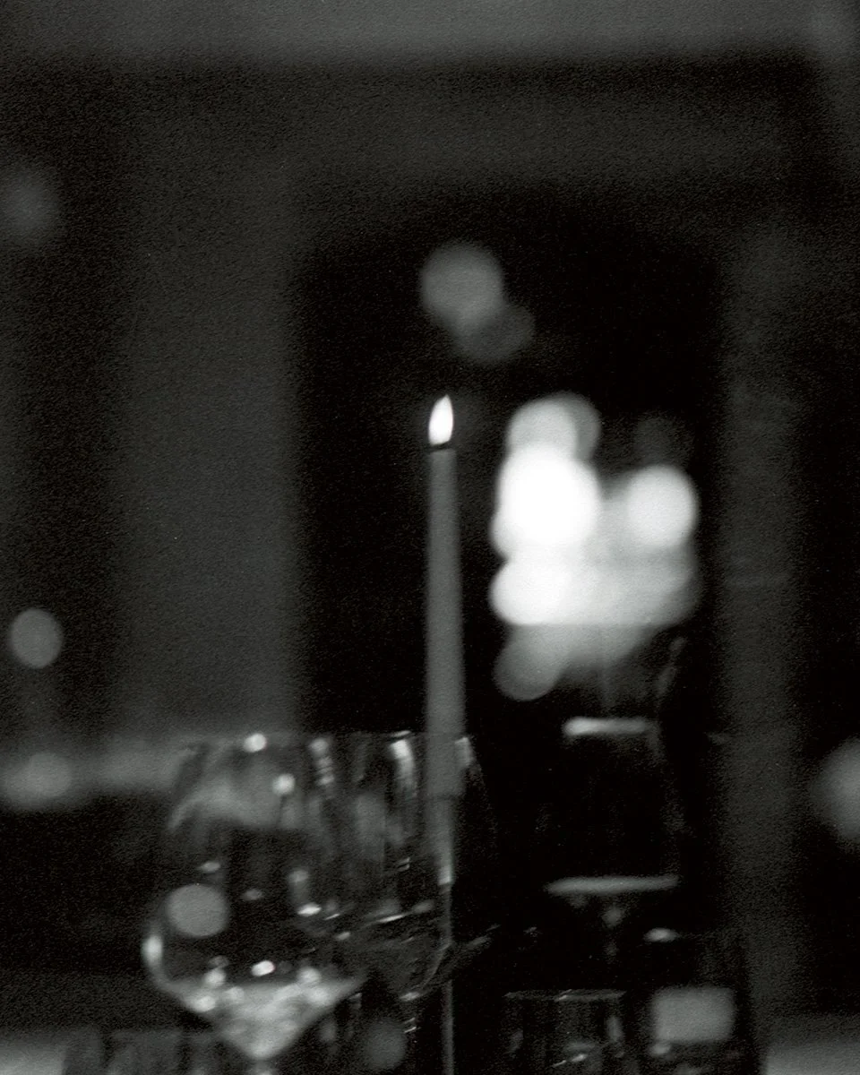 A lit candle on a table with wine glasses and blurred lights in the background, in black and white.