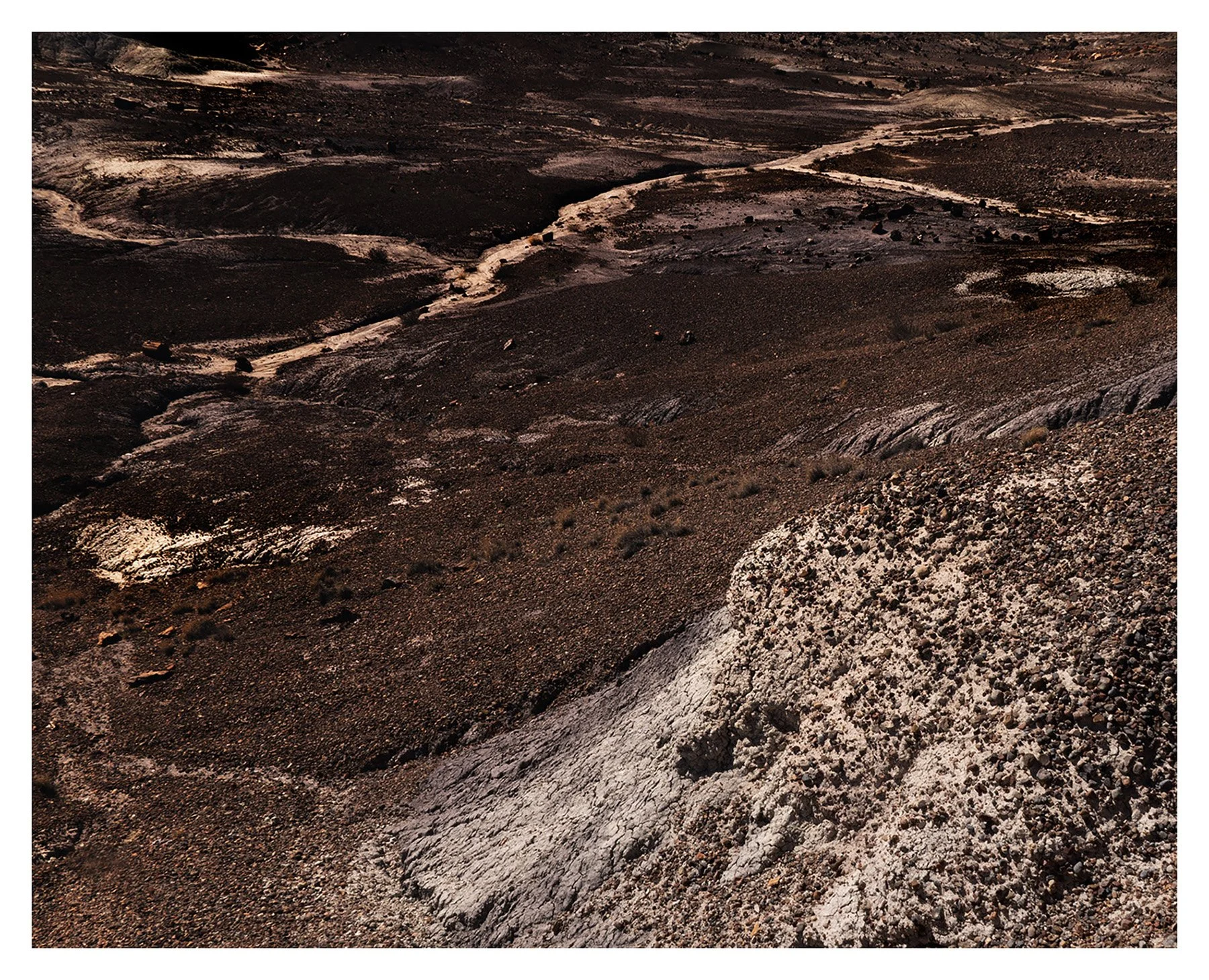 A barren, rocky landscape with dark soil and light cracks, featuring winding dirt paths and scattered rocks, with no visible vegetation or structures.
