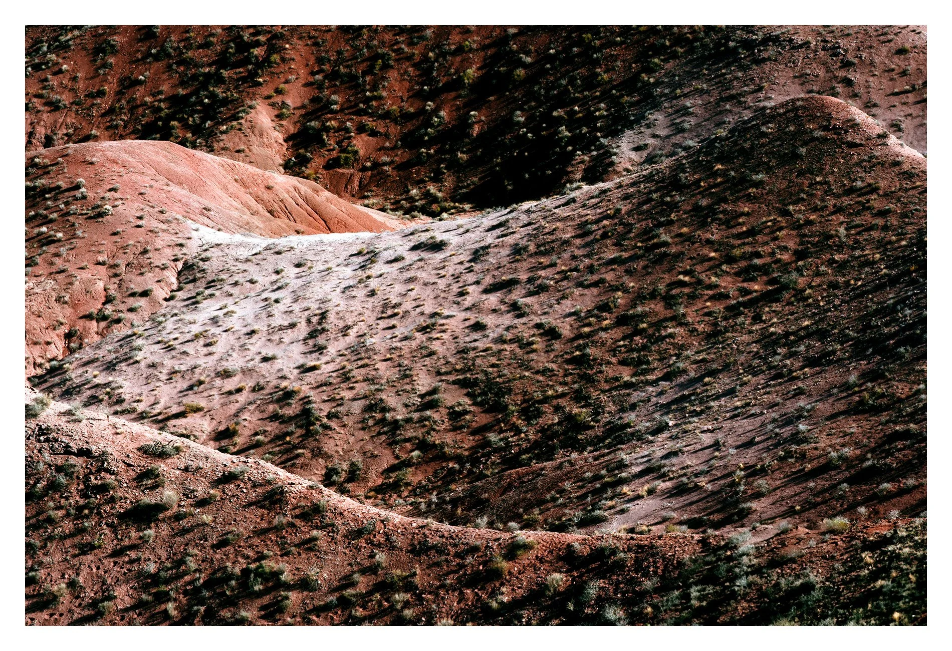 Aerial view of desert hills with reddish-brown soil and sparse vegetation, creating a pattern of shadows and highlights across the landscape.