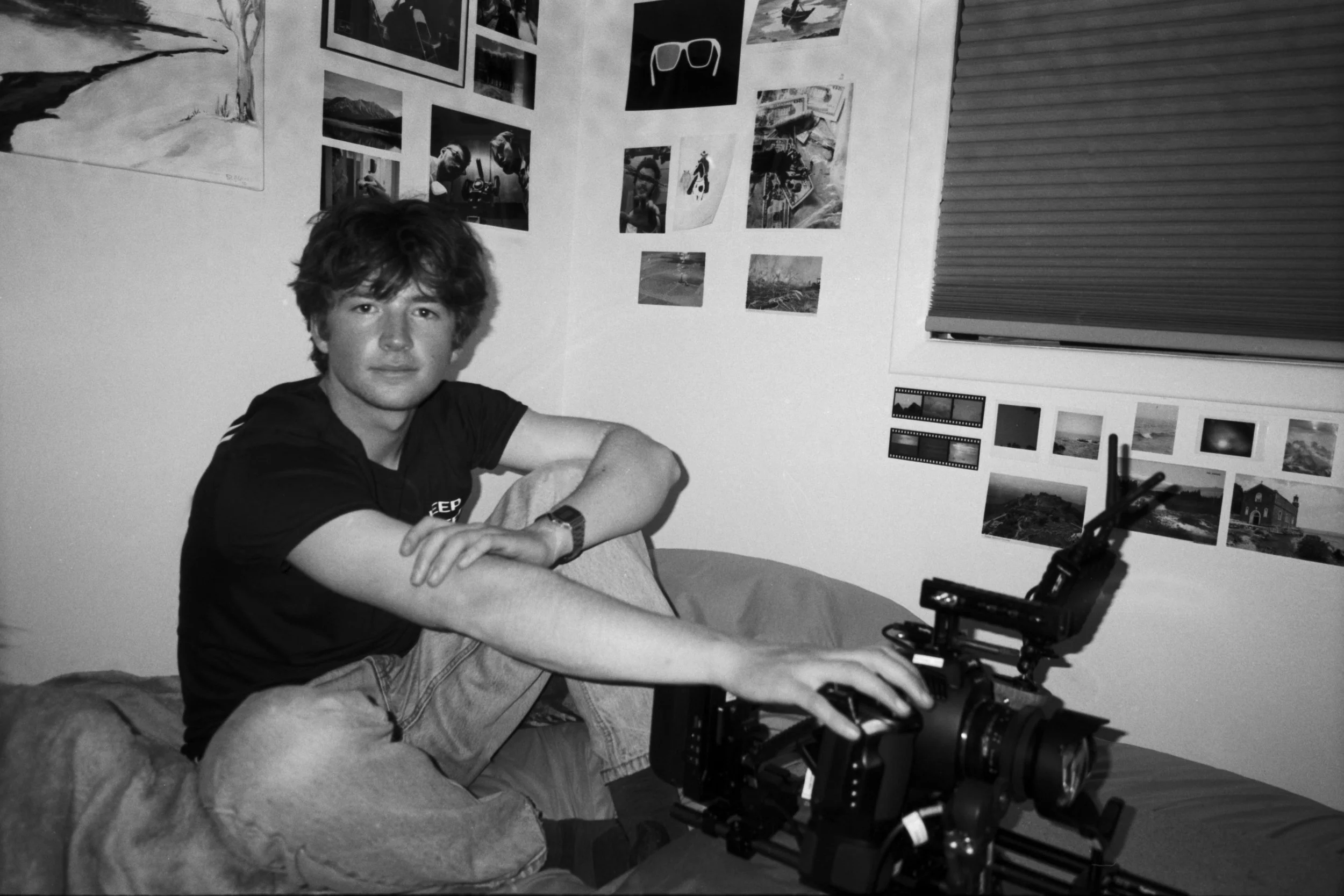 A young man with shaggy hair sitting on a bed in a room with walls decorated with landscape and travel photographs, operating a camera mounted on a tripod.